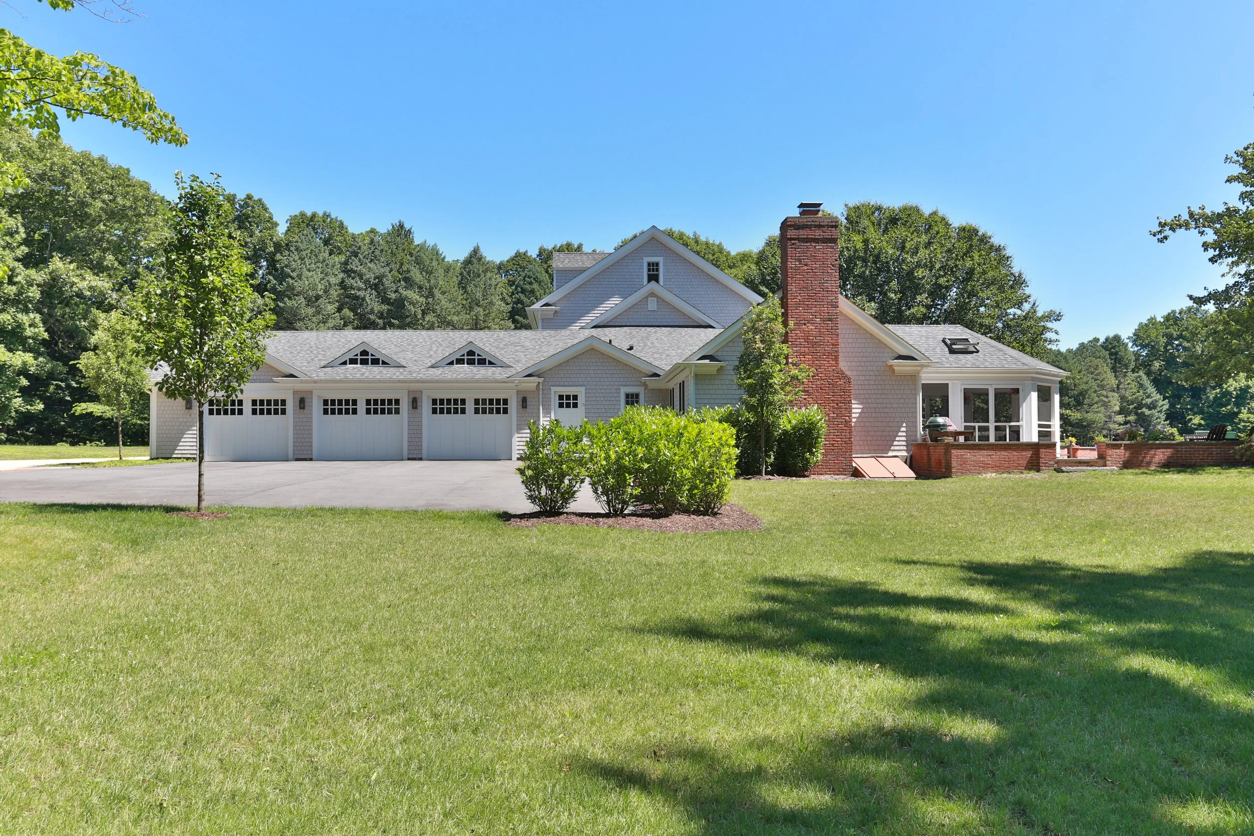 Front view of a large house with a three-car garage, a brick chimney, and a porch, surrounded by green lawn and trees under a clear blue sky.