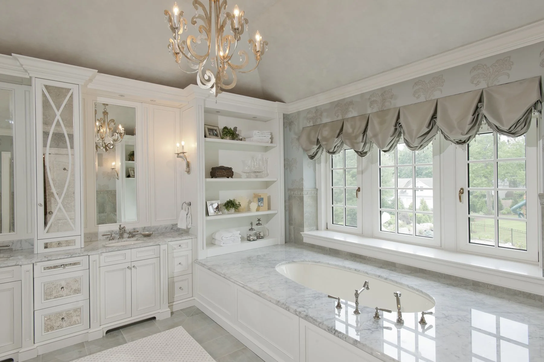 Luxury bathroom with large window, marble bathtub, built-in shelves, elegant chandelier, and ornate window treatments.