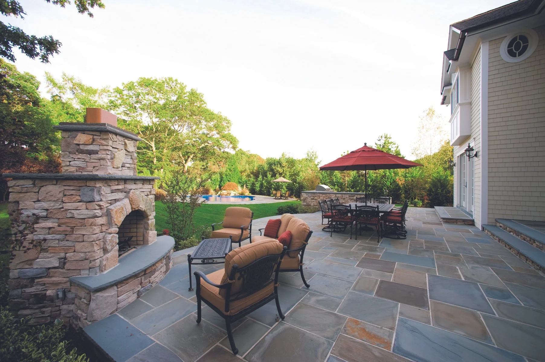 Backyard patio with stone fireplace, outdoor furniture, umbrella, grass yard with trees, in-ground pool, and house siding.