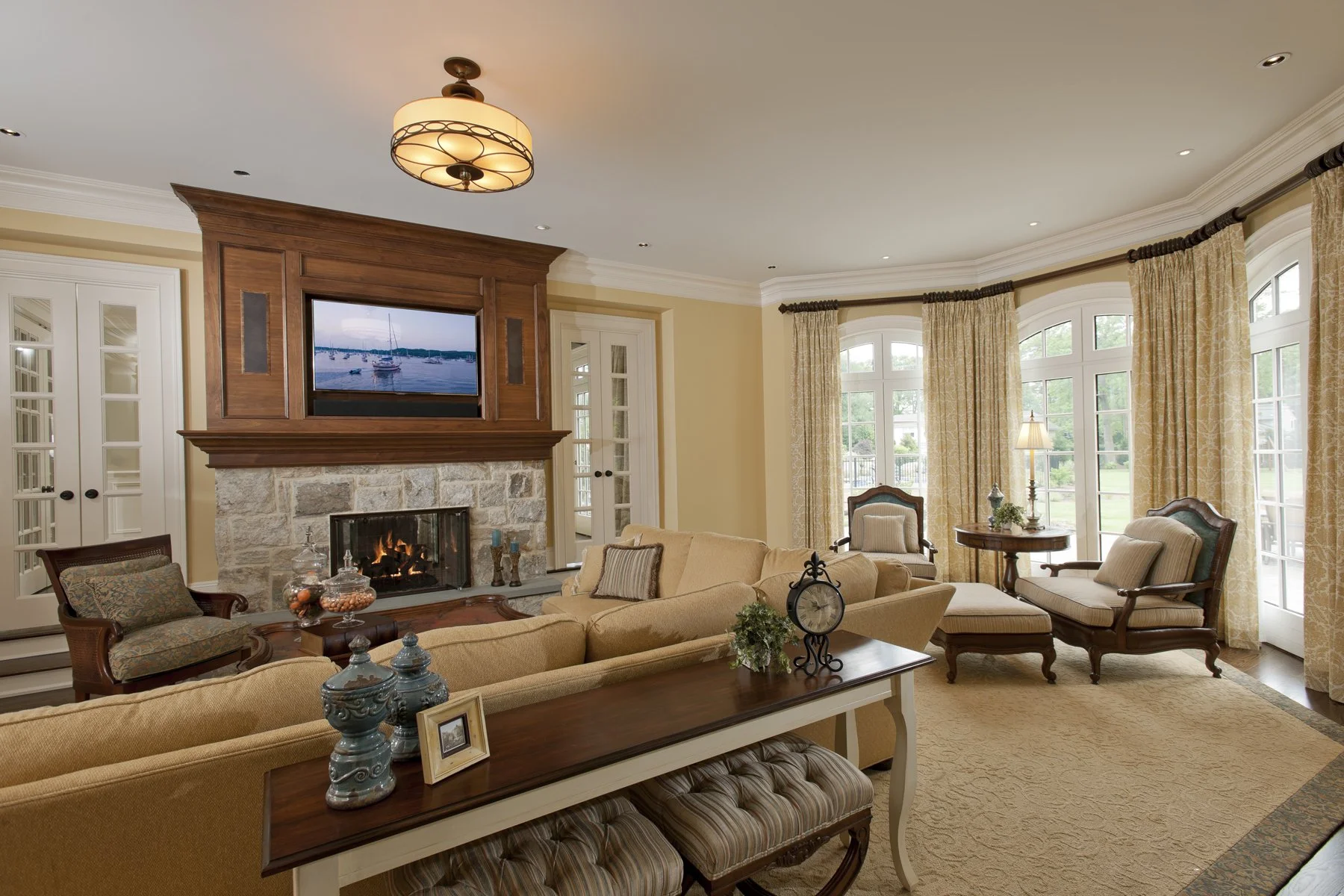 Living room with fireplace, stone mantel, and large windows with beige curtains; beige sofas, wooden armchairs, and a round table with a lamp