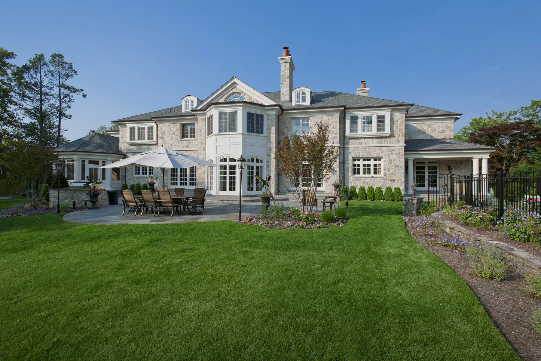 Large stone house with multiple chimneys, a patio with outdoor dining table and umbrella, well-maintained lawn, and garden with flowers, trees, and a black iron fence.