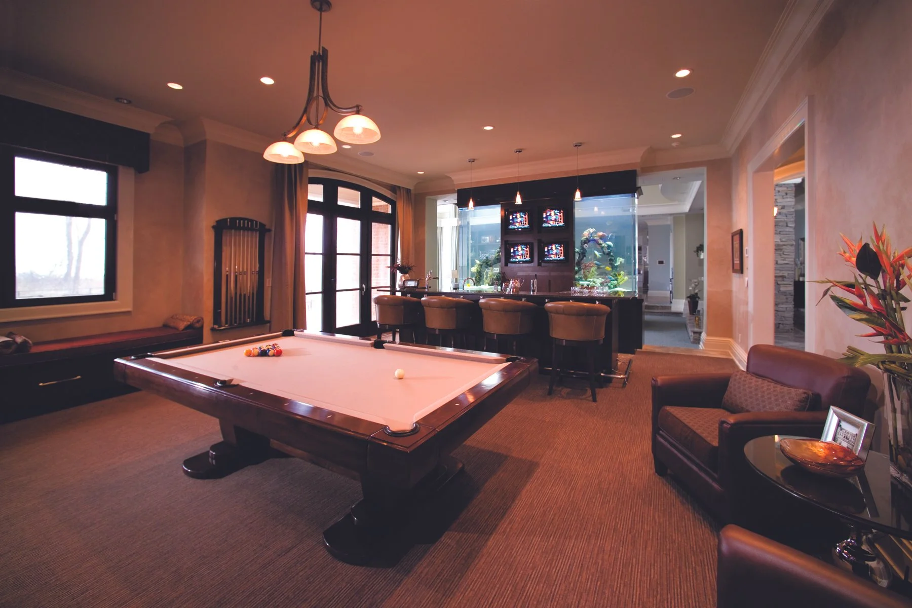 A cozy game room with a pool table, bar area with barstools, an aquarium, and comfortable chairs, illuminated by warm lighting.