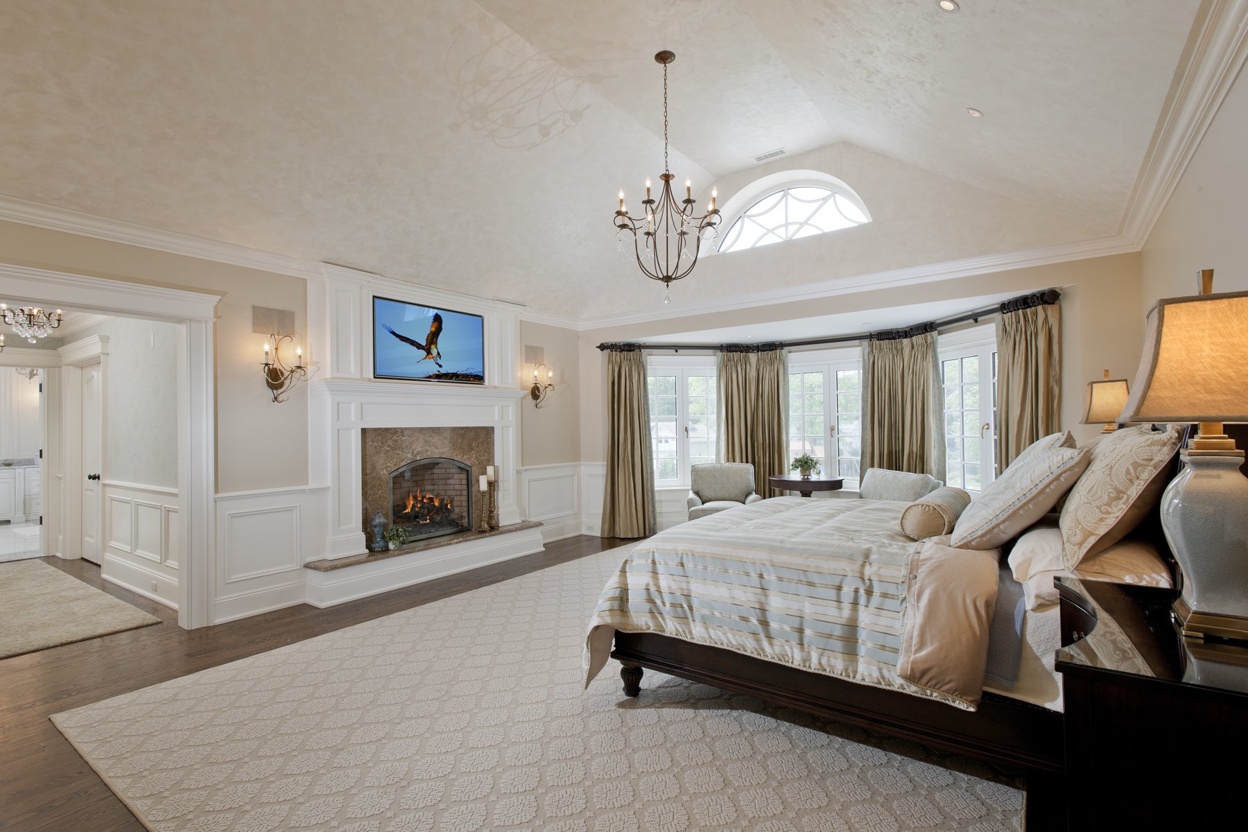 Elegant bedroom with a large bed, a fireplace, a wall-mounted TV, and large bay windows with curtains.