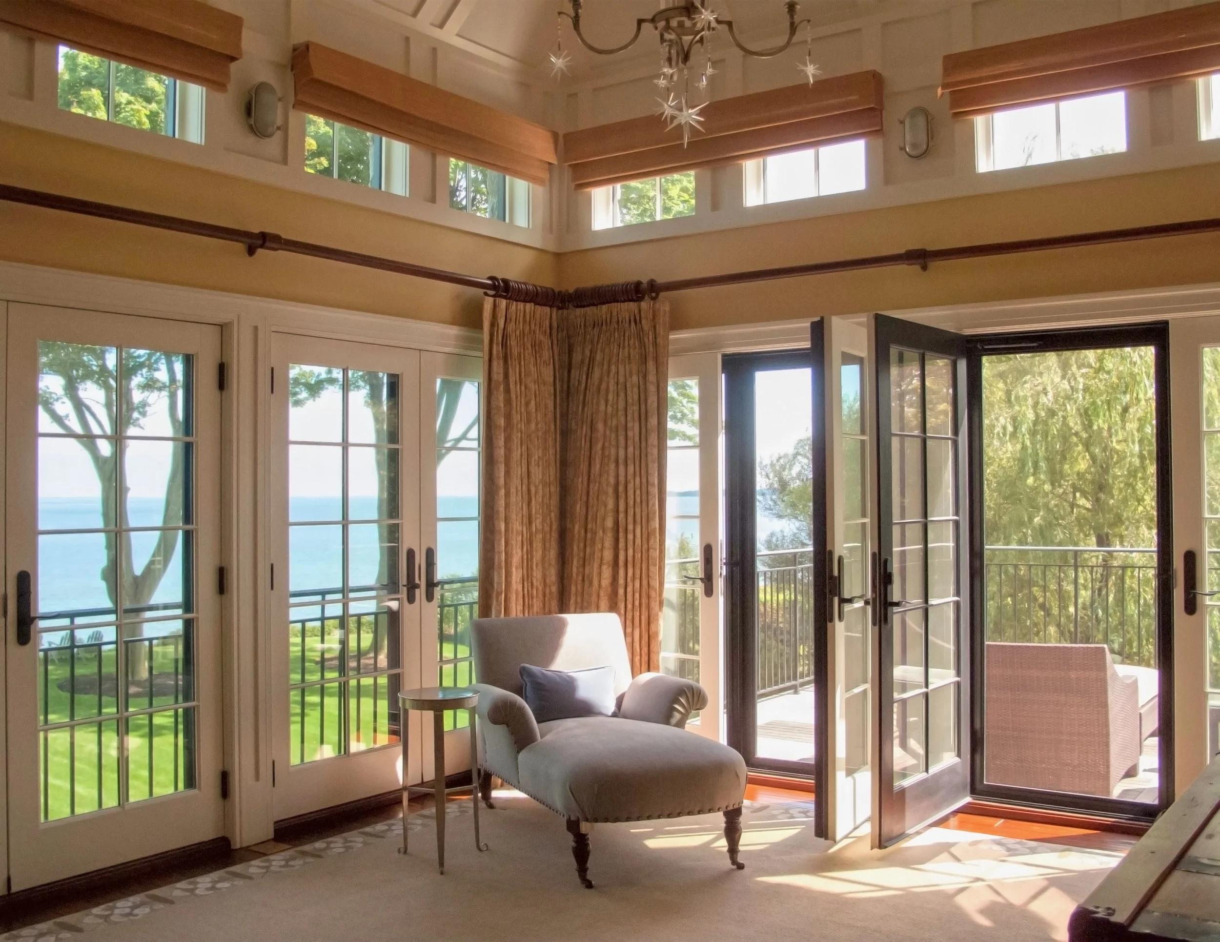 Sunlit living room with large glass doors opening to a balcony overlooking water and greenery, featuring an armchair, side table, and view of trees and outdoors.