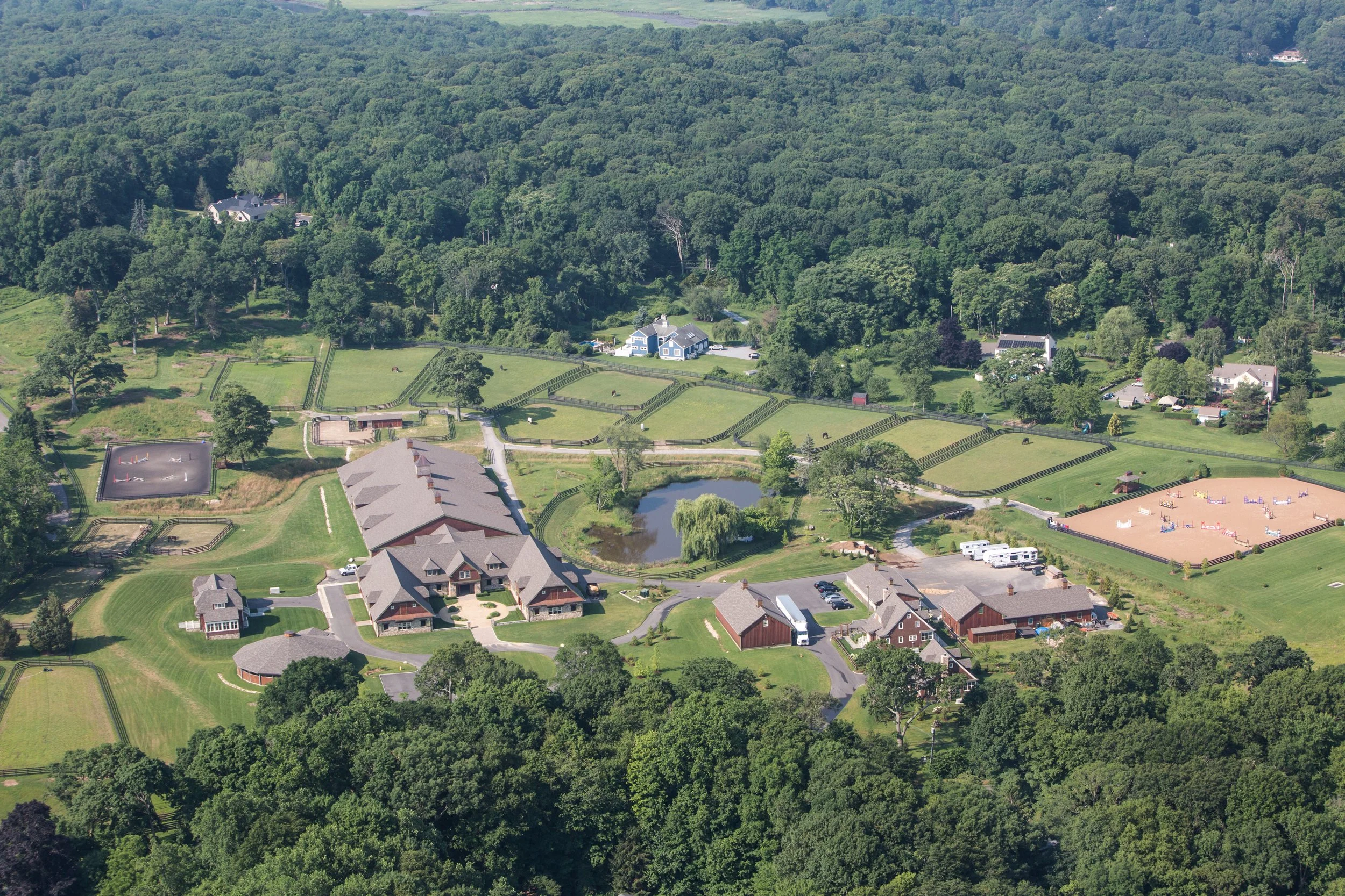 A large countryside estate with multiple buildings, a pond, tennis courts, and horse riding arenas surrounded by trees and greenery.