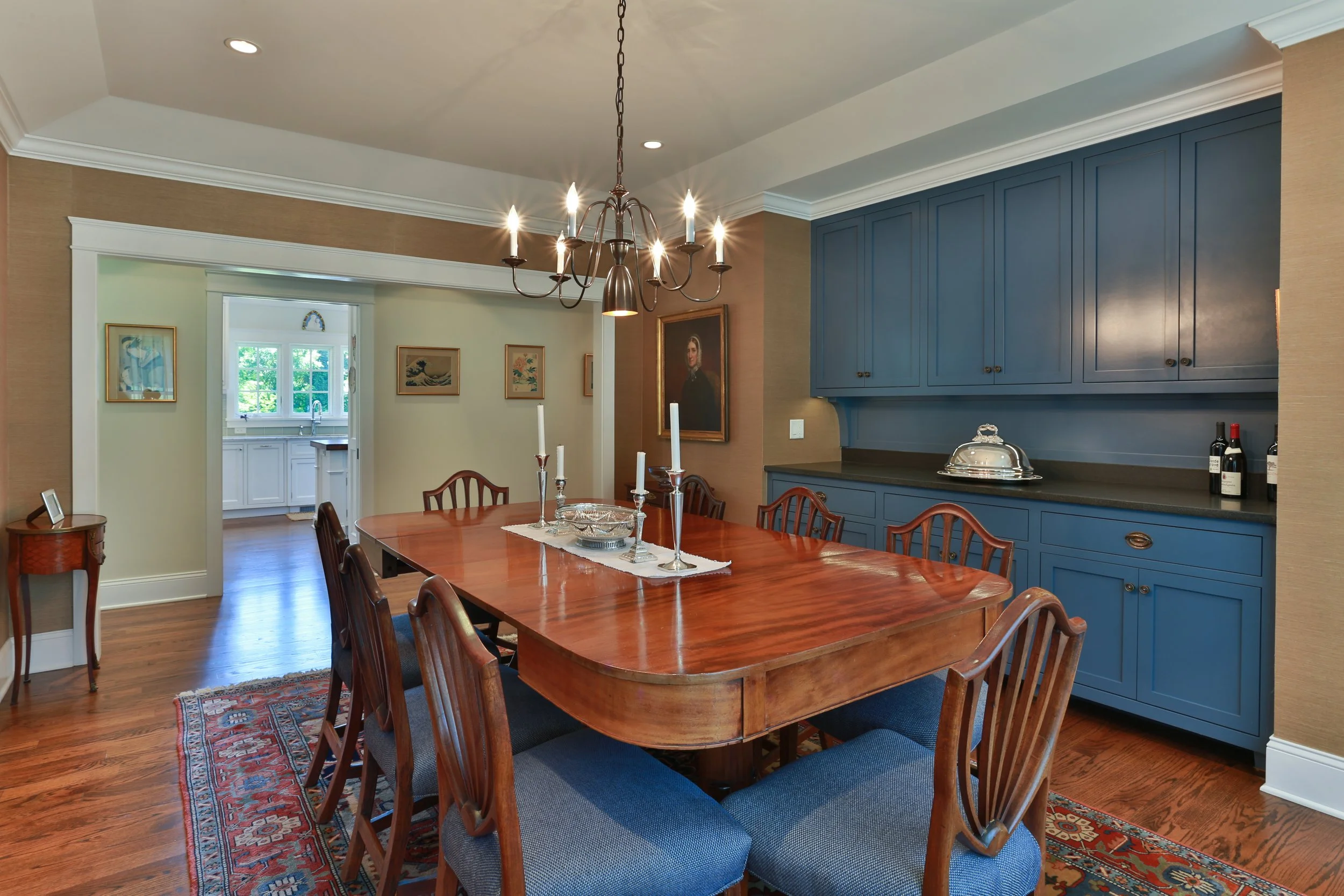 A dining room with a wooden table, blue upholstered chairs, a chandelier, and blue cabinets against the wall. There are candles and wine bottles on the sideboard. An entrance leads to a bright kitchen with white cabinets.