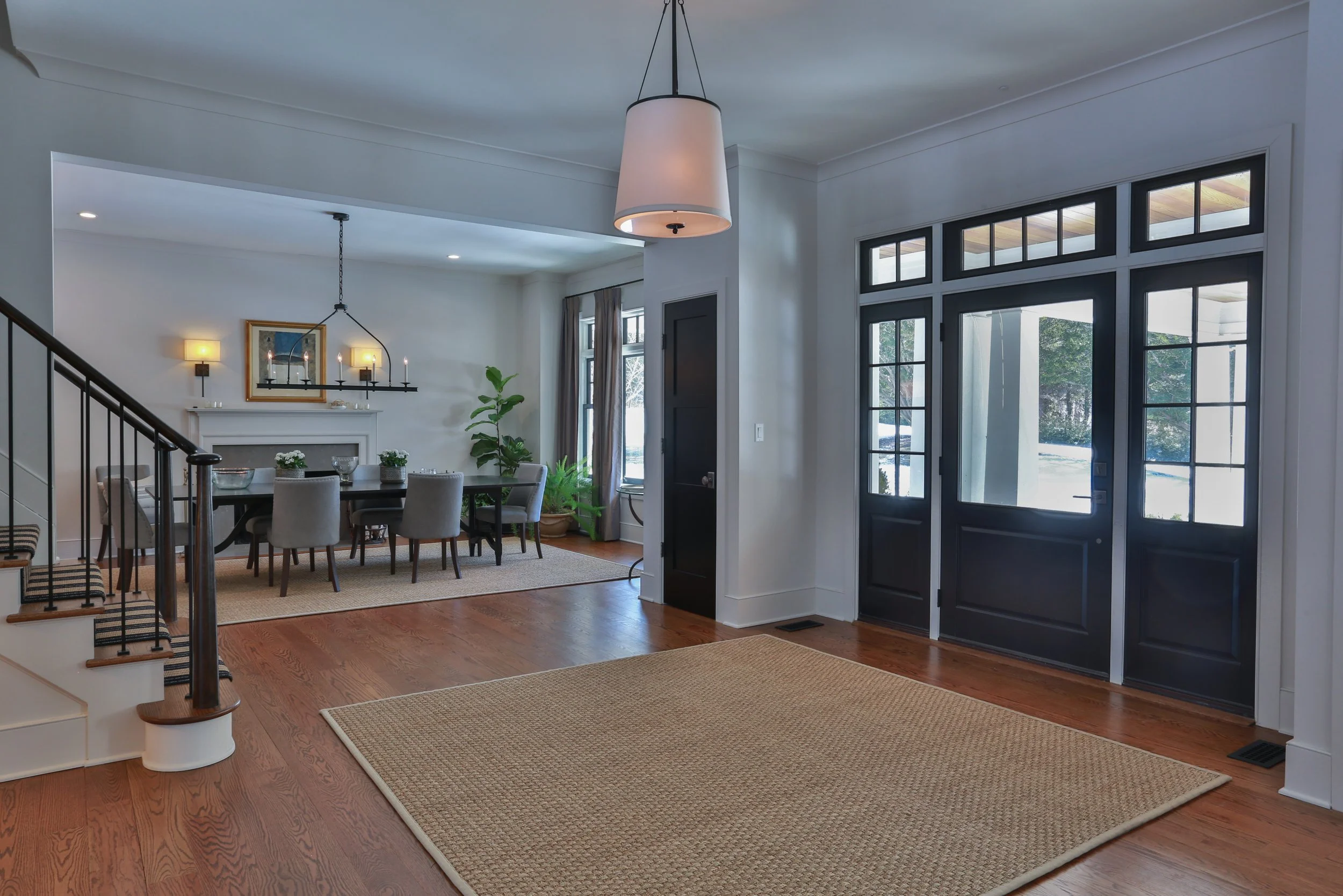 Interior view of a modern house with a wooden floor, black front door and large windows, leading into a dining area with a table, gray chairs, a black chandelier, and wall-mounted lights.
