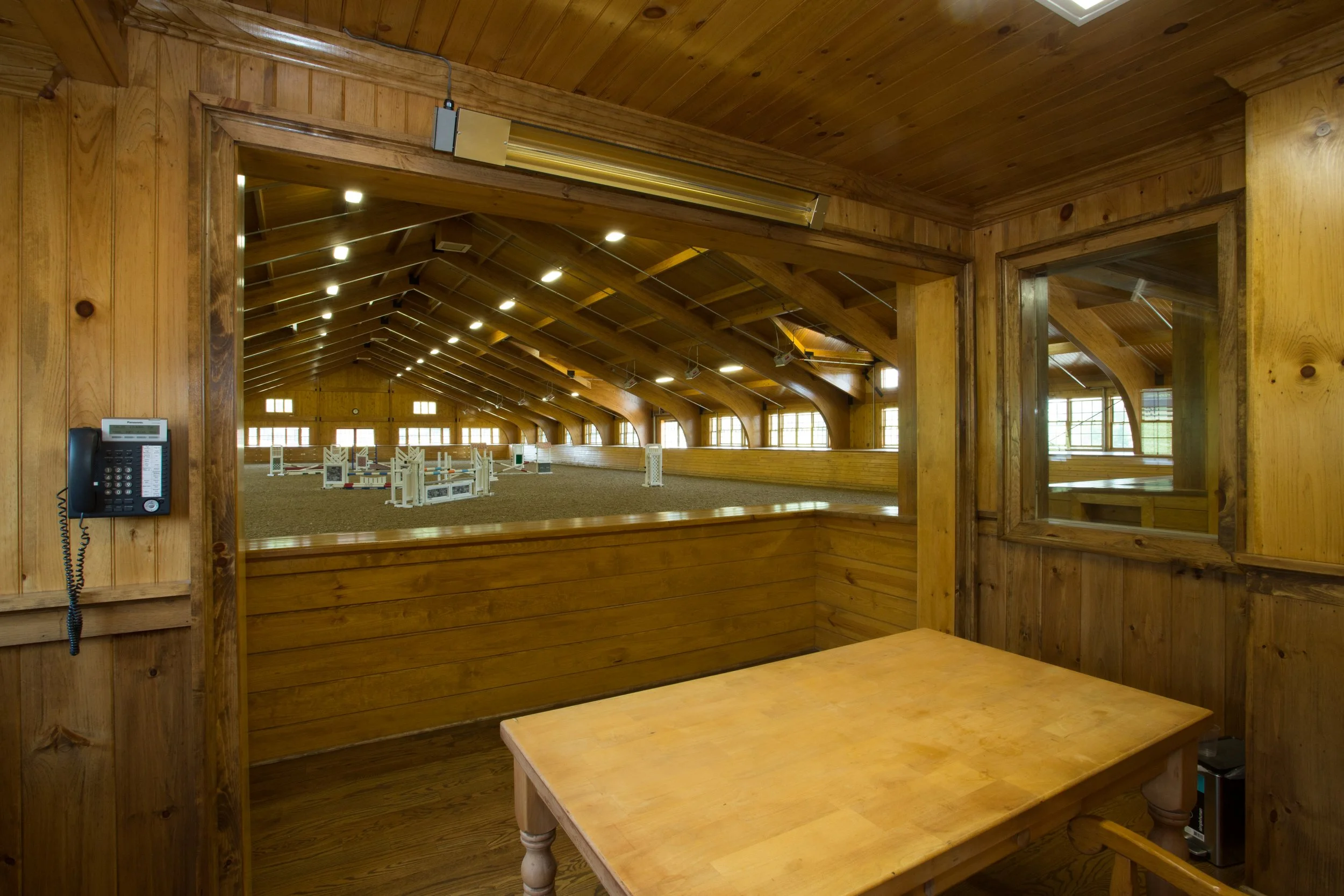 View from a wooden enclosed room overlooking an indoor horse riding arena with jumping obstacles.
