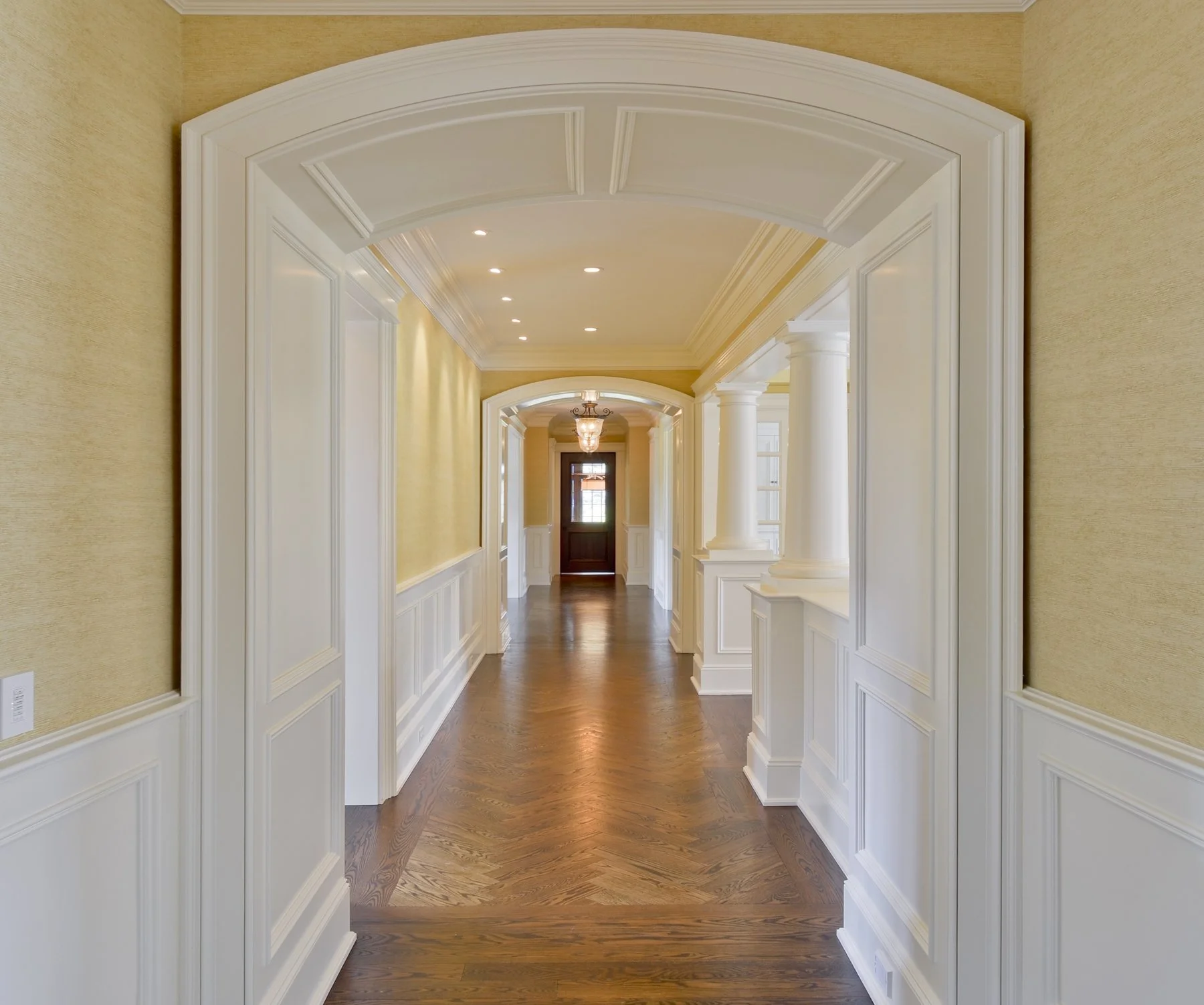 Bright hallway with hardwood floors, cream-colored walls, white wainscoting, and decorative molding. There are multiple ceiling lights and a chandelier near the dark front door in the background.