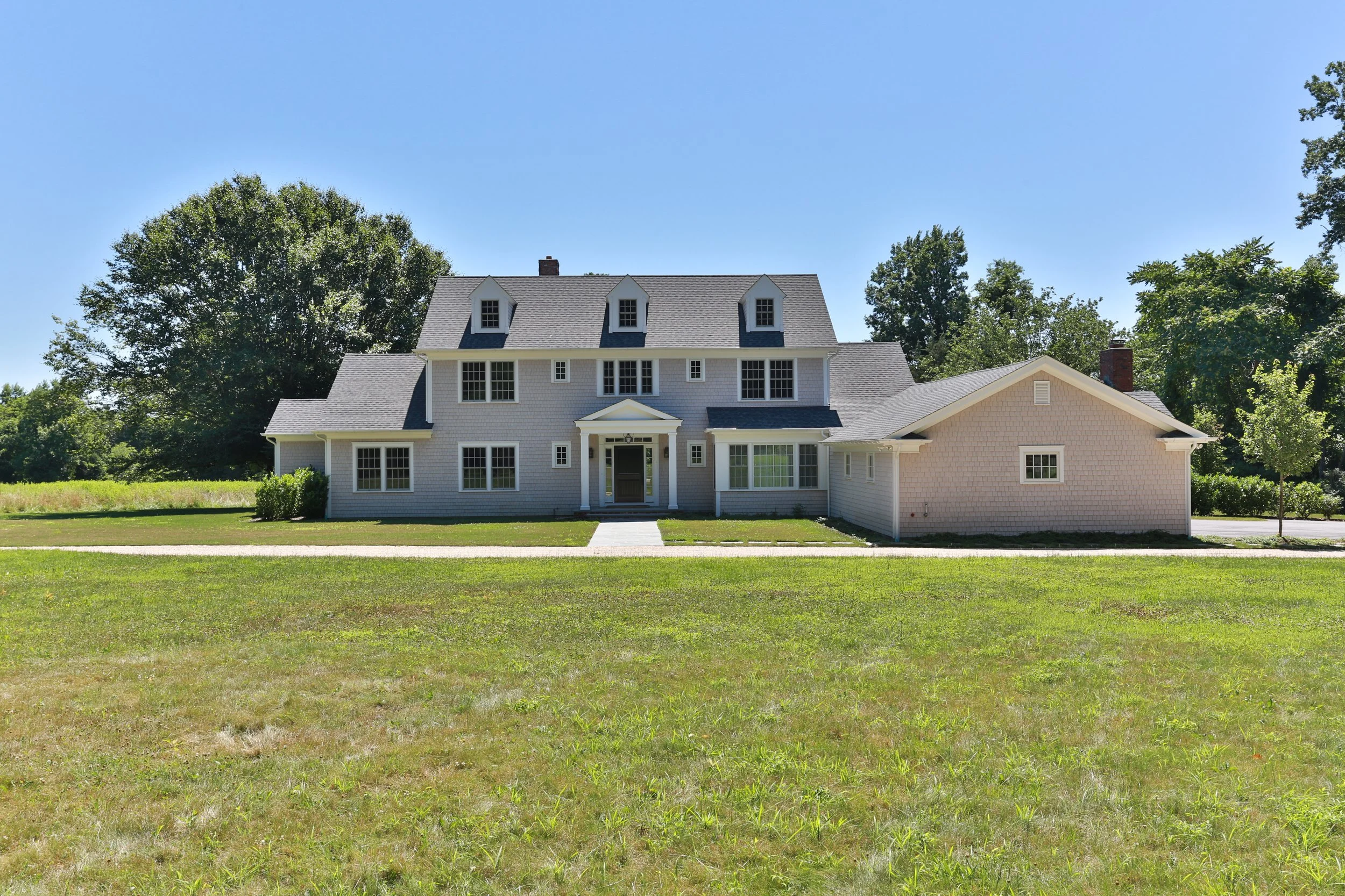 Front view of a large, white, two-story house with multiple dormer windows, a front porch, and a well-kept lawn, surrounded by trees under a clear blue sky.