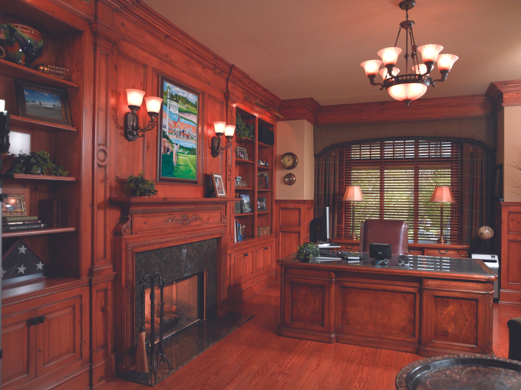 A luxurious office with wood-paneled walls and furniture, a fireplace, a large window with blinds and curtains, a desk with a chair, and decorative lighting.