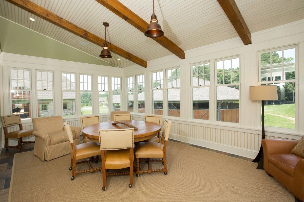 Sunroom with large windows, wooden beams on white ceiling, round wooden dining table, six upholstered chairs, beige carpet, leather armchair, floor lamp, and outside views of houses and green lawn