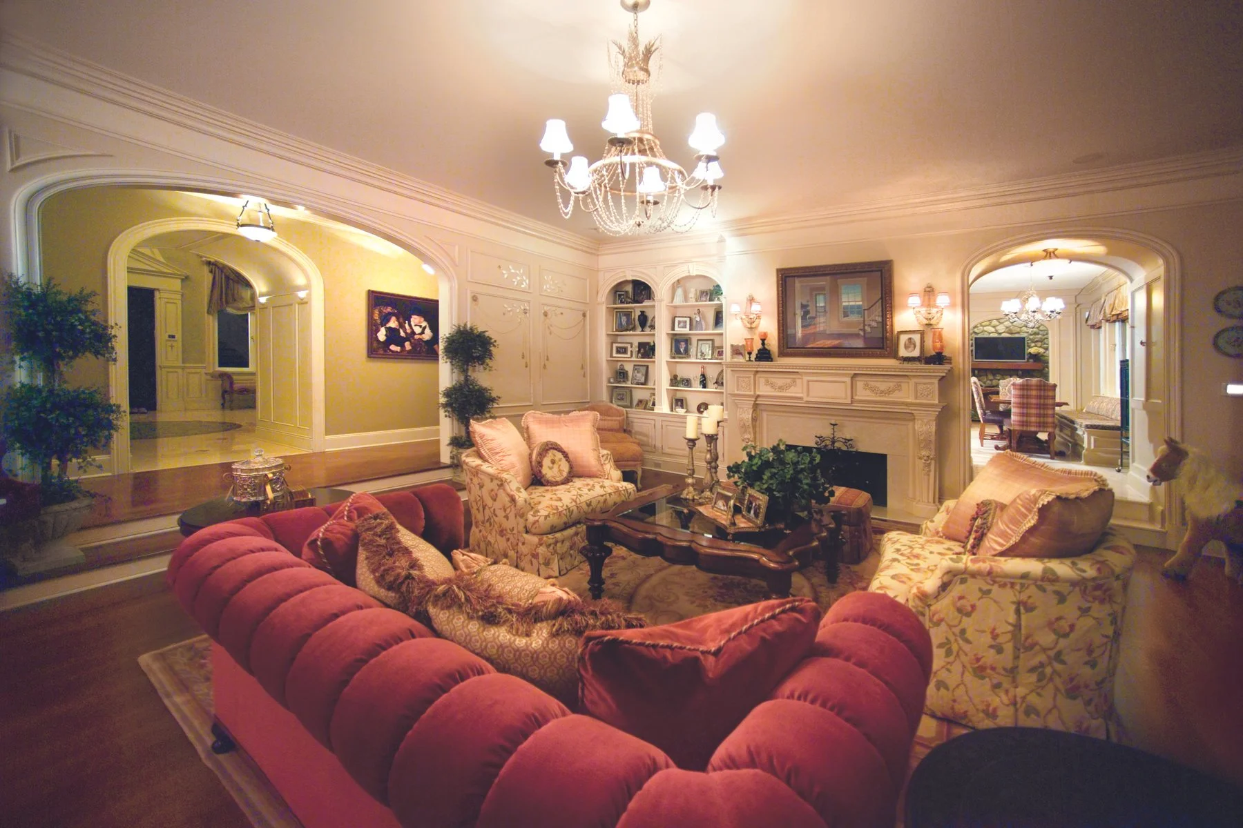 Elegant living room with a red velvet sofa, floral armchairs, a dark wood coffee table, a fireplace, and decorative lighting.