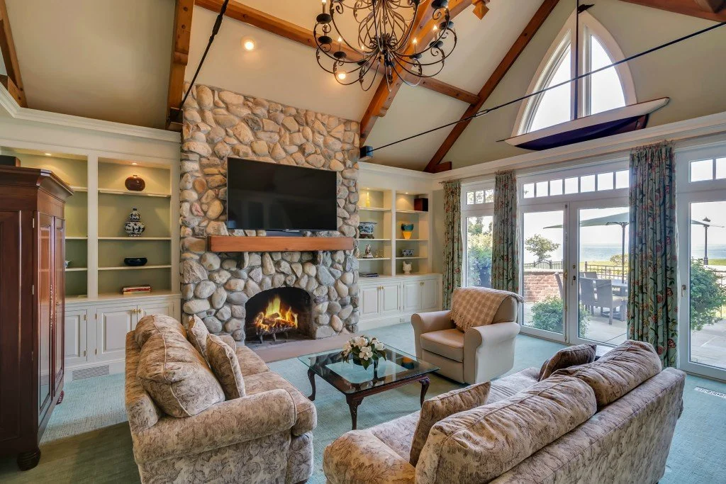 Living room with stone fireplace, mounted flat-screen TV, beige sofas, armchair, glass coffee table, and sliding glass doors leading to a patio with outdoor furniture and ocean view.