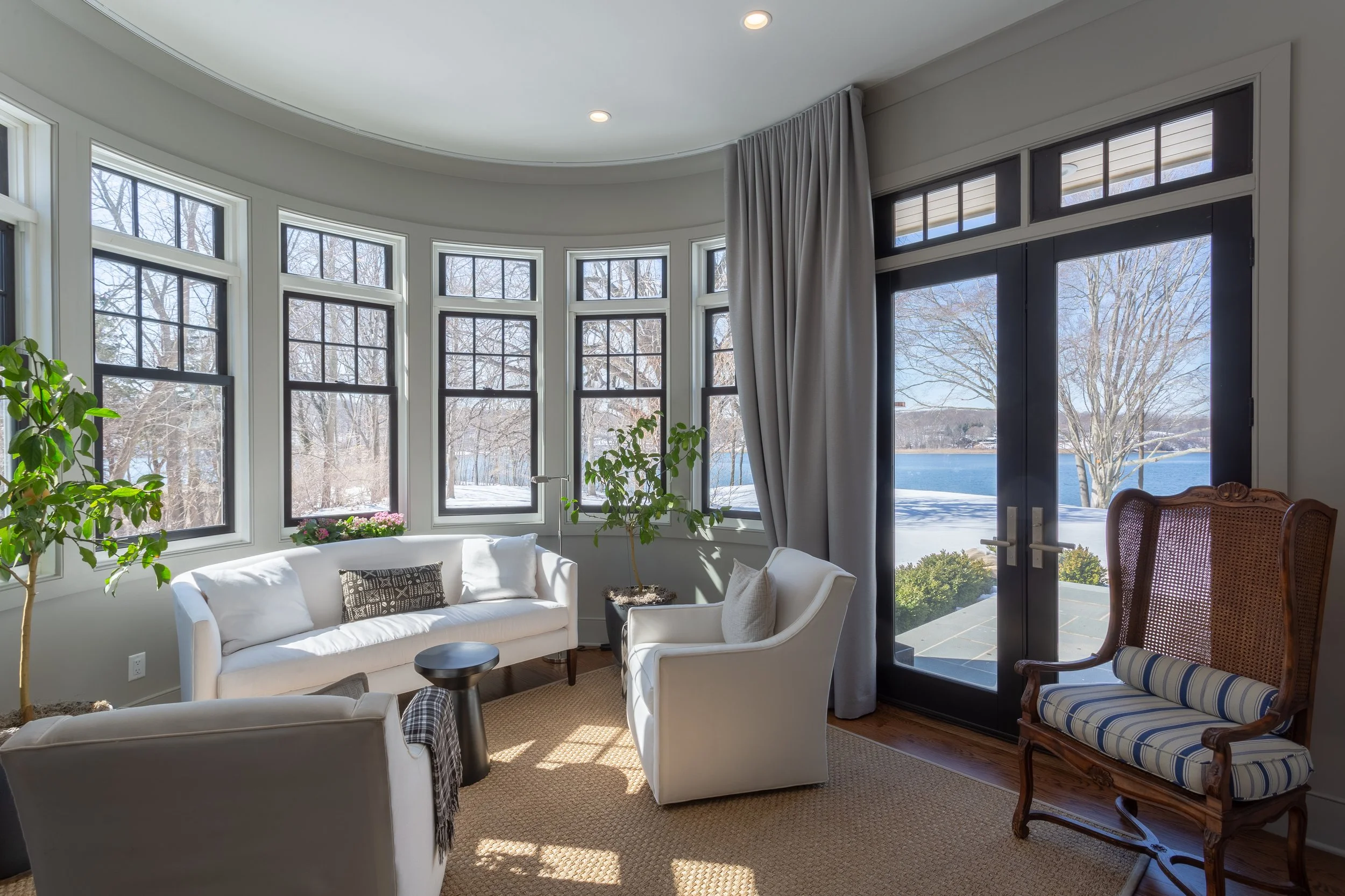 Living room with large bay windows and glass doors, overlooking a snowy yard and lake, furnished with white sofas, a wooden chair with a striped cushion, and potted plants.