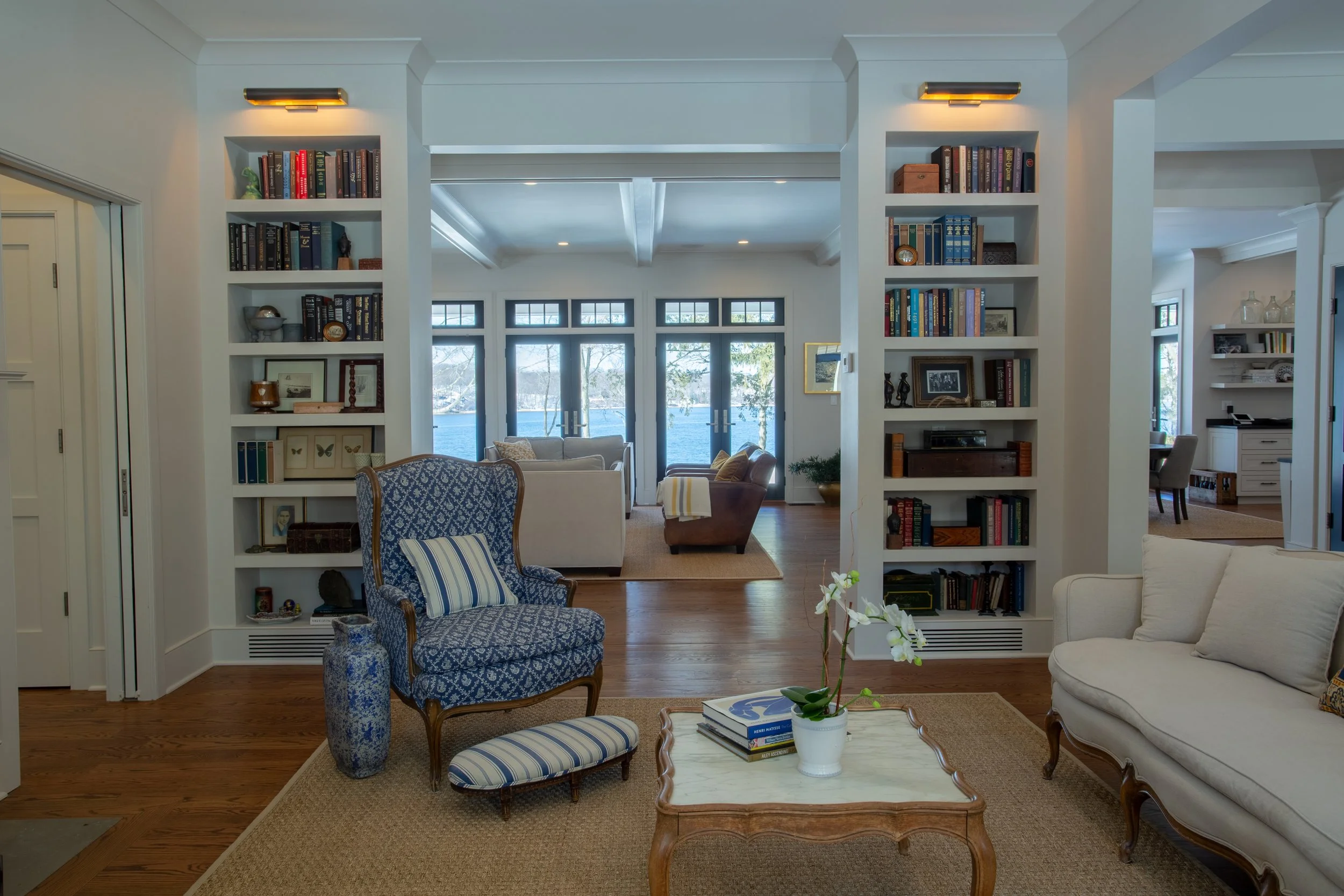 Living room with built-in bookshelves, hardwood floors, and large windows overlooking a lake, furnished with sofas, armchairs, and a coffee table with a potted orchid.