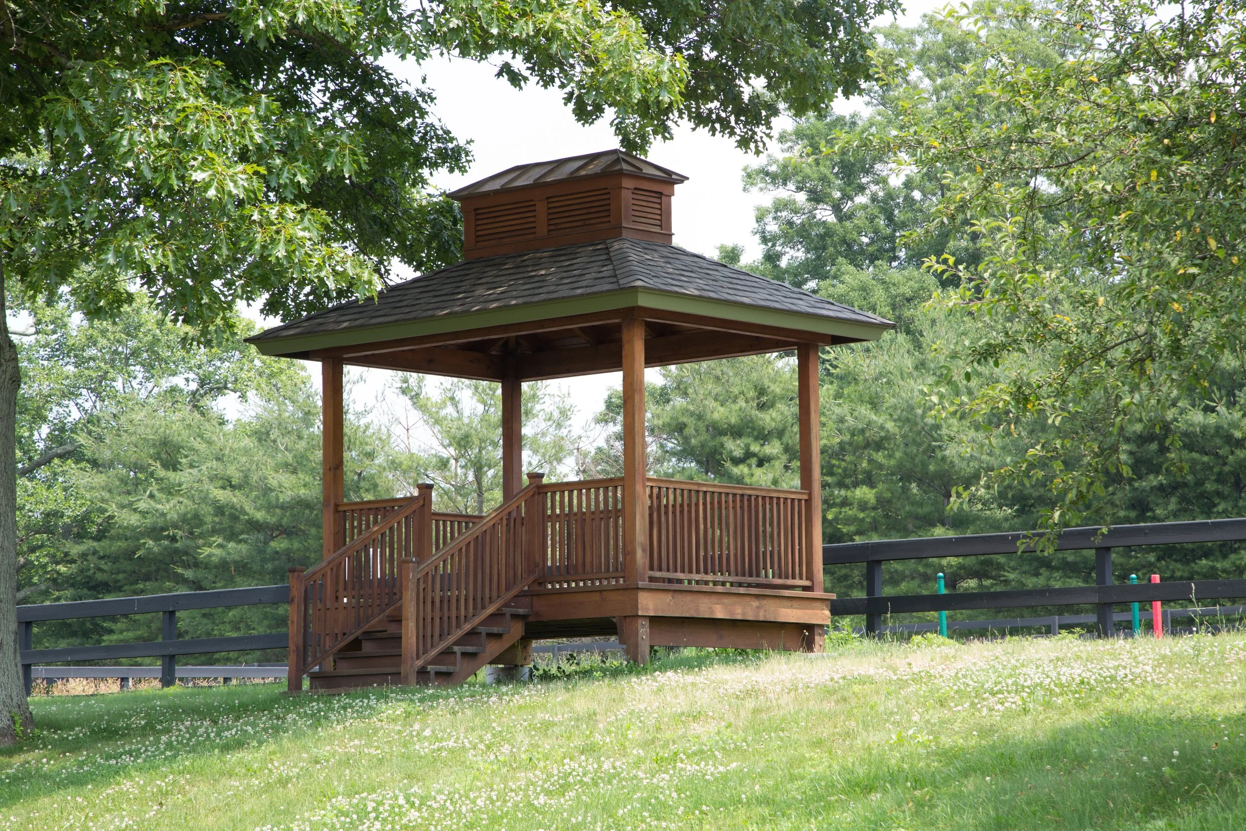 Wooden gazebo with stairs, surrounded by green grass and trees, with a black fence in the background.