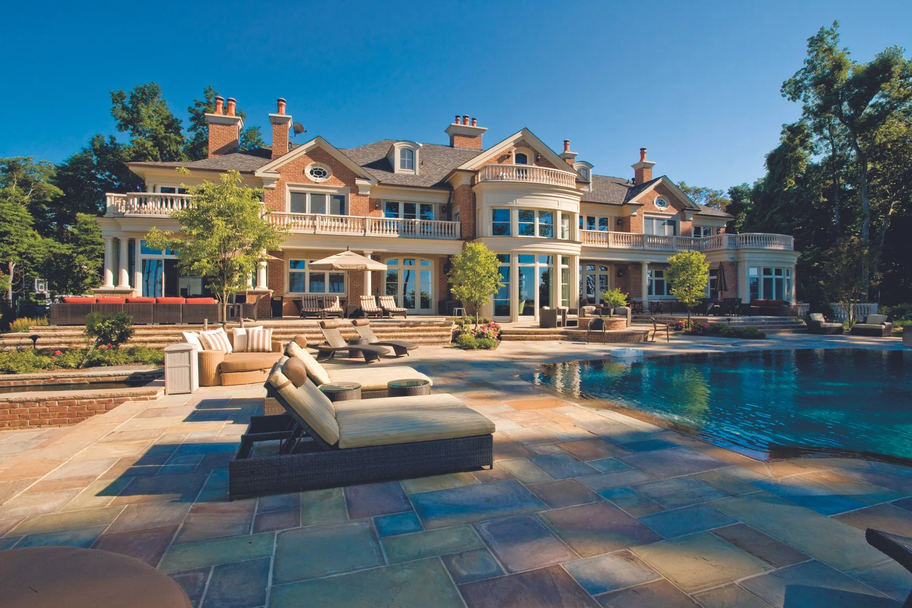 Large luxurious house with multiple stories, large windows, and balconies, situated behind a pool area with lounge chairs, umbrellas, and lush greenery, under a clear blue sky.