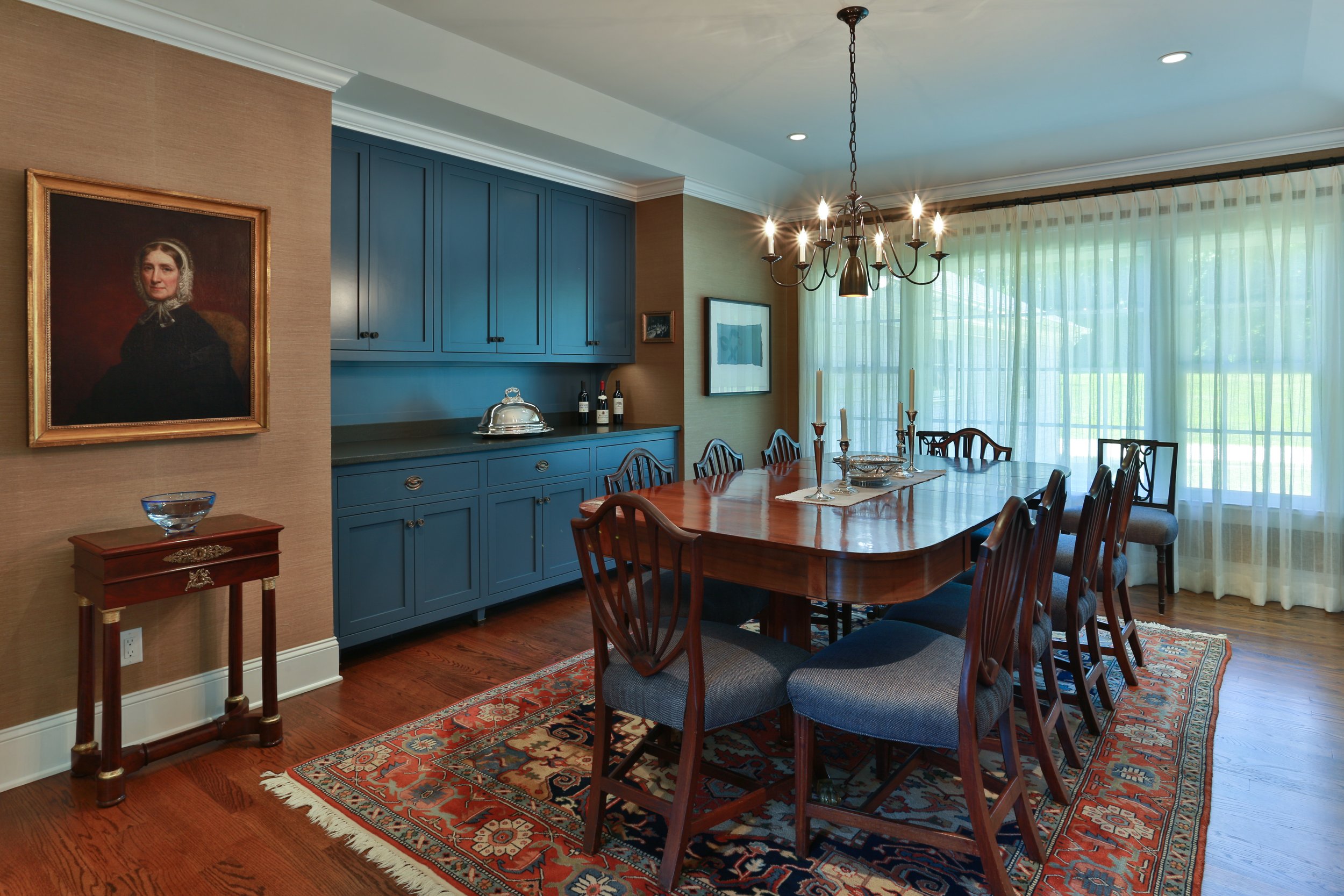 A dining room with a large wooden table surrounded by ten chairs, a chandelier hanging above, and a large window with sheer curtains letting in natural light.
