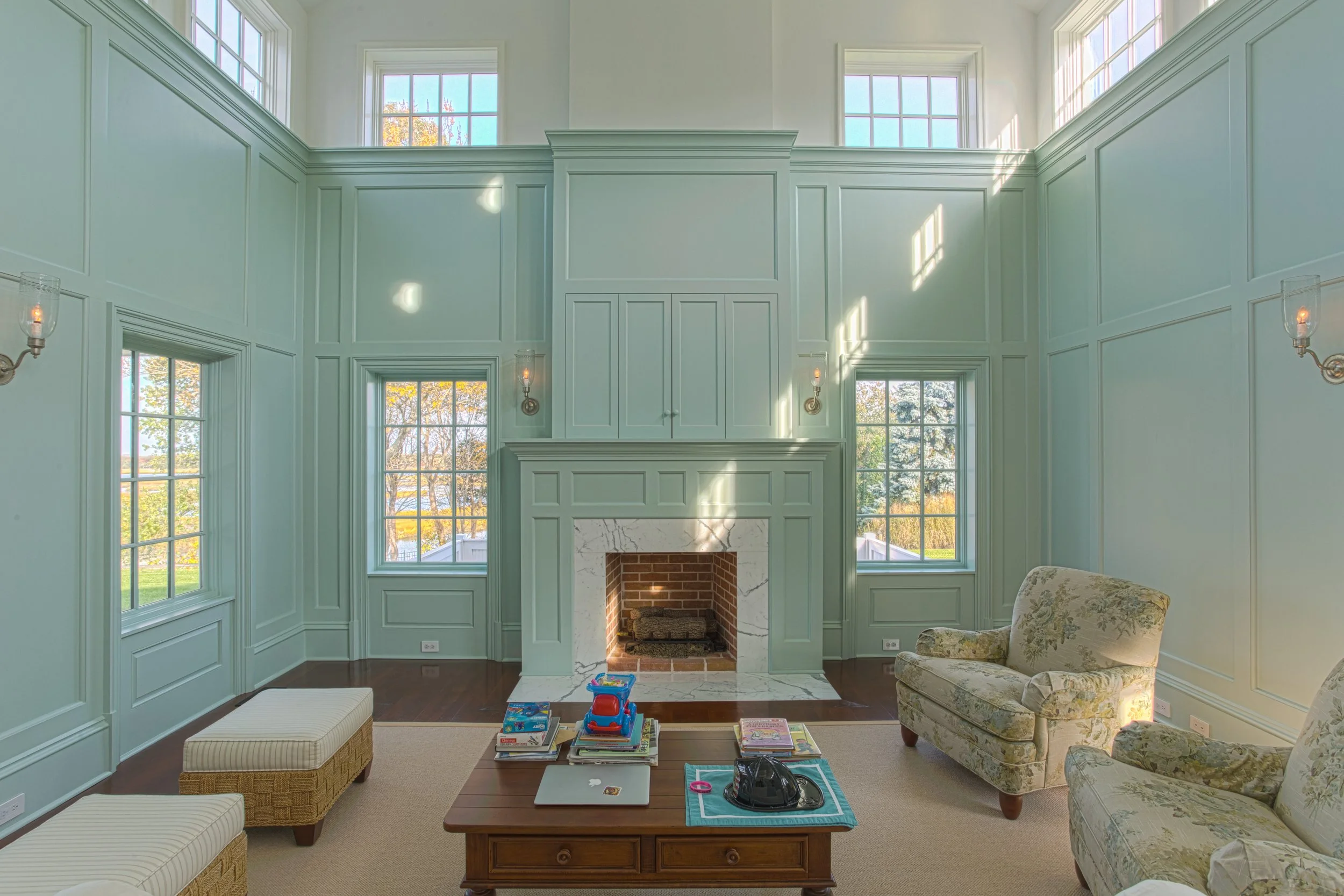 Living room with pastel green paneled walls, large windows letting in natural light, a white marble and brick fireplace, floral upholstered armchairs, a wooden coffee table with toys and magazines, and a beige carpet.
