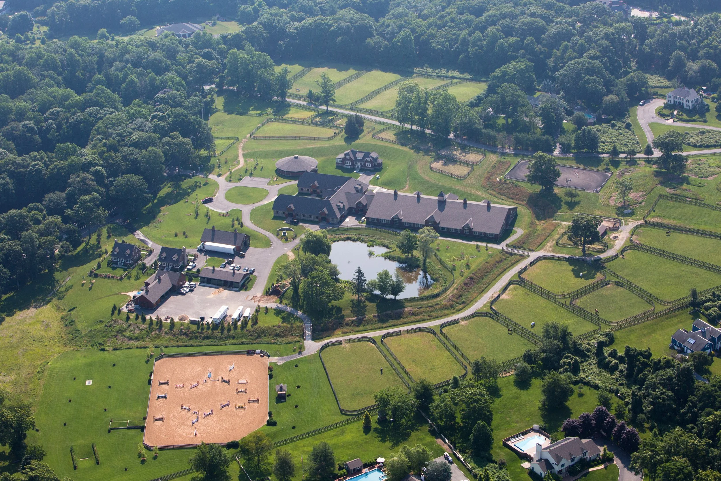 Aerial view of a large estate with multiple buildings, a pond, tennis courts, riding arenas, and fenced grassy areas, surrounded by trees.