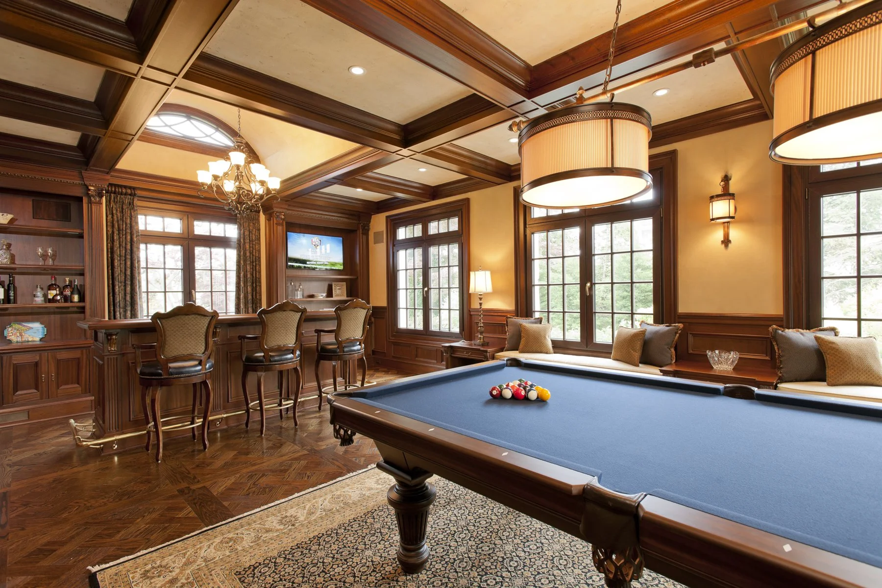 Elegant game room with wooden paneling, a blue pool table, windows, bar area with chairs, flat-screen TV, chandelier, pendant lights, and seating with pillows.