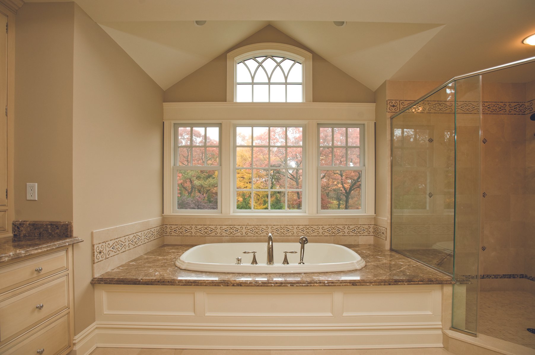 Bathroom with a large window view of trees with fall foliage, a bathtub on a marble platform, and a glass shower.