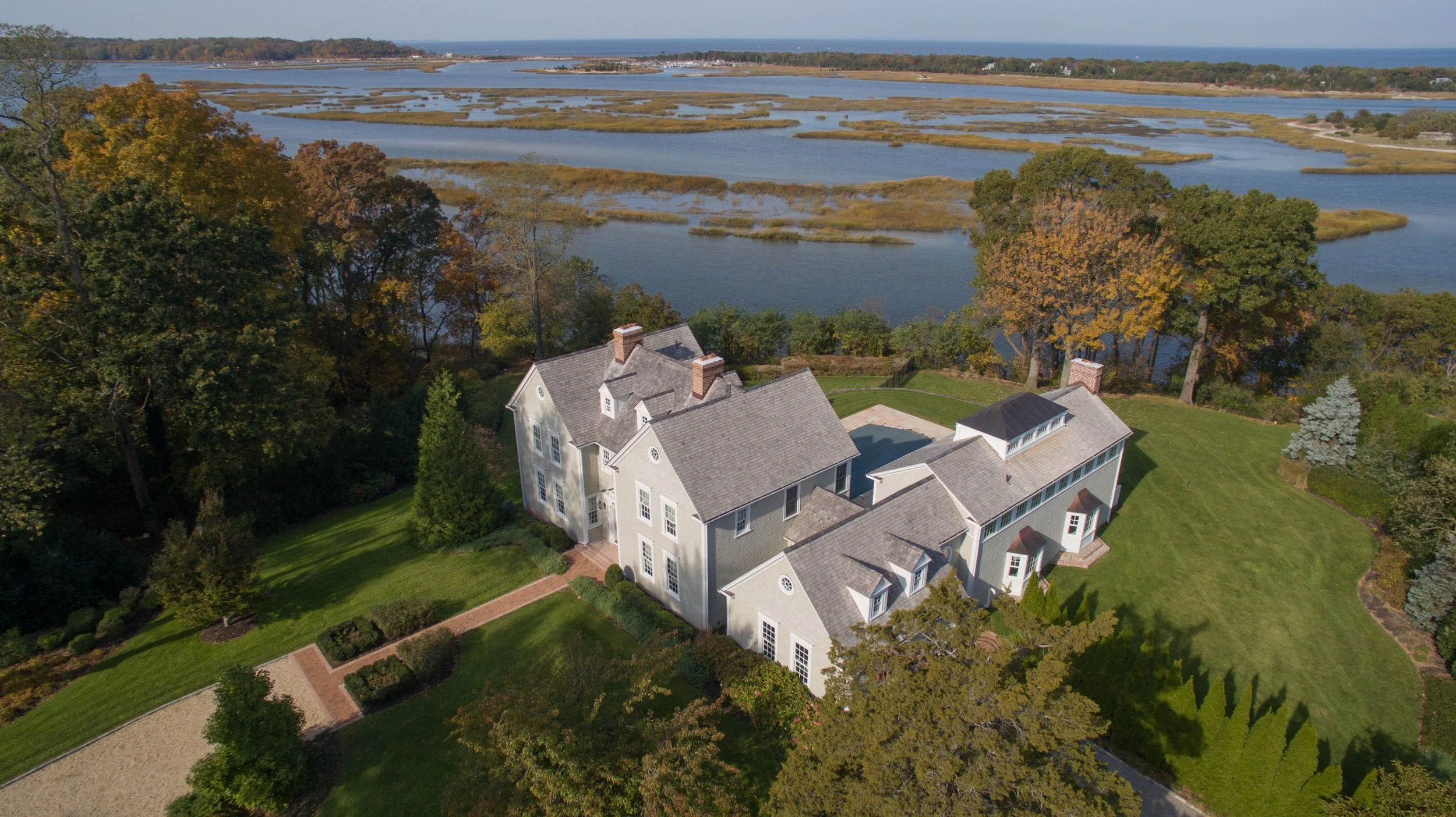 Aerial view of a large, white mansion with a gray roof surrounded by green lawn and trees, overlooking a body of water with marshes and islands in the background.