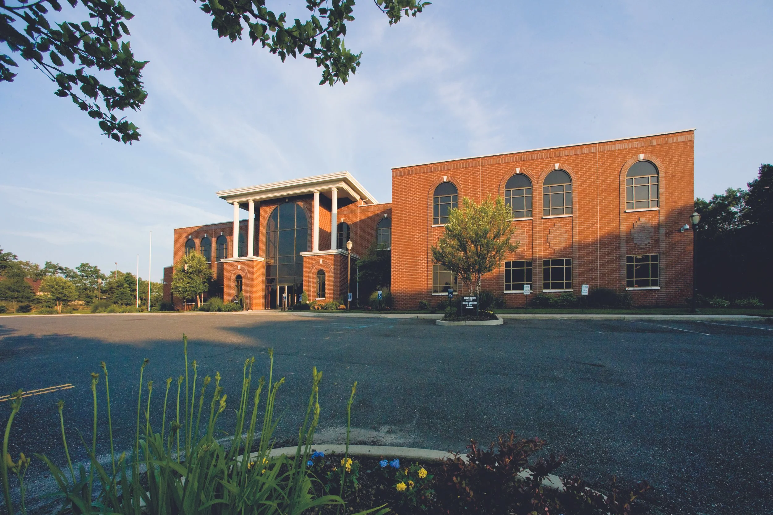 A large brick building with many windows and a prominent entrance with white columns, surrounded by a parking lot with several handicap parking spaces, trees, and shrubs.