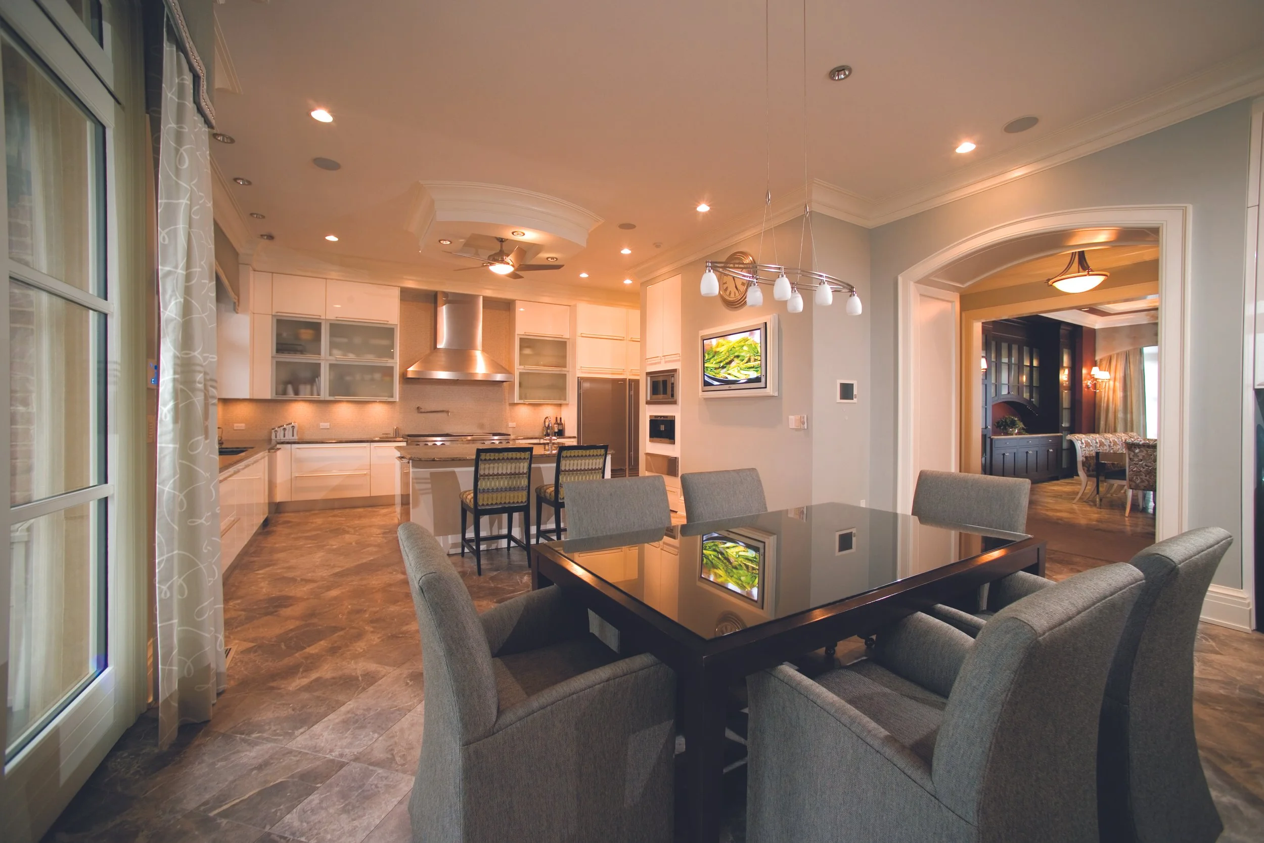 Open-concept living space with a dining area and kitchen, modern decor, gray chairs around a dark wood table, bright lighting, and a view into a formal sitting room in the background.