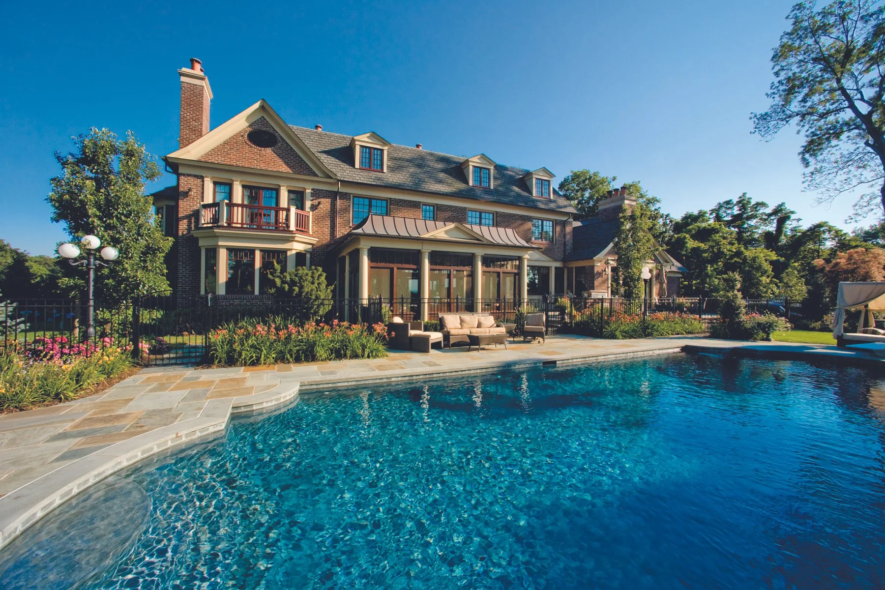 Large brick house with multiple gabled roofs and dormer windows, surrounded by green trees and landscaped gardens, with a swimming pool in the foreground.