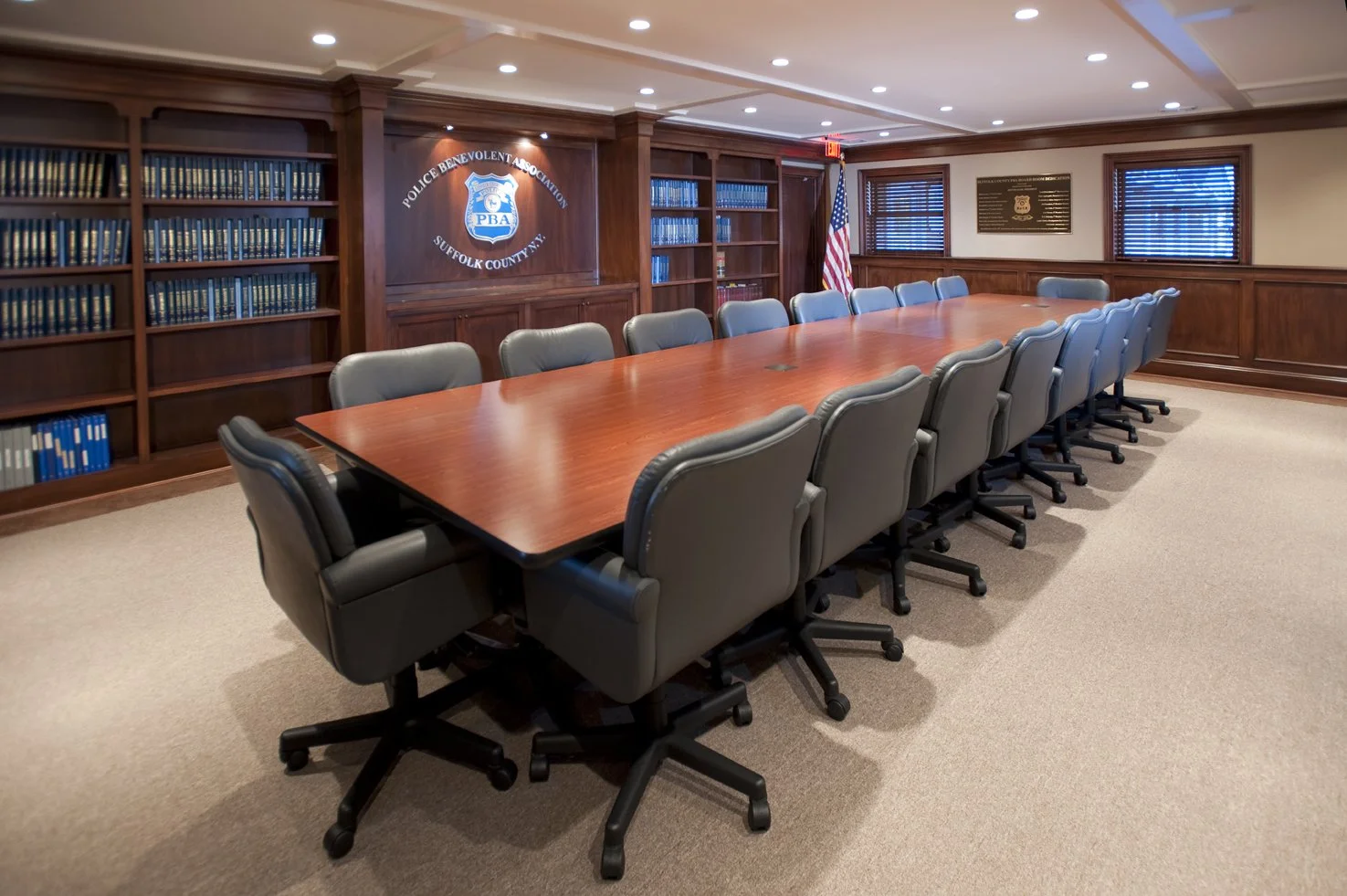A conference room with a long wooden table surrounded by black leather office chairs, wooden bookshelves with blue binders, a United States flag, and a sign indicating it is a police association meeting room.