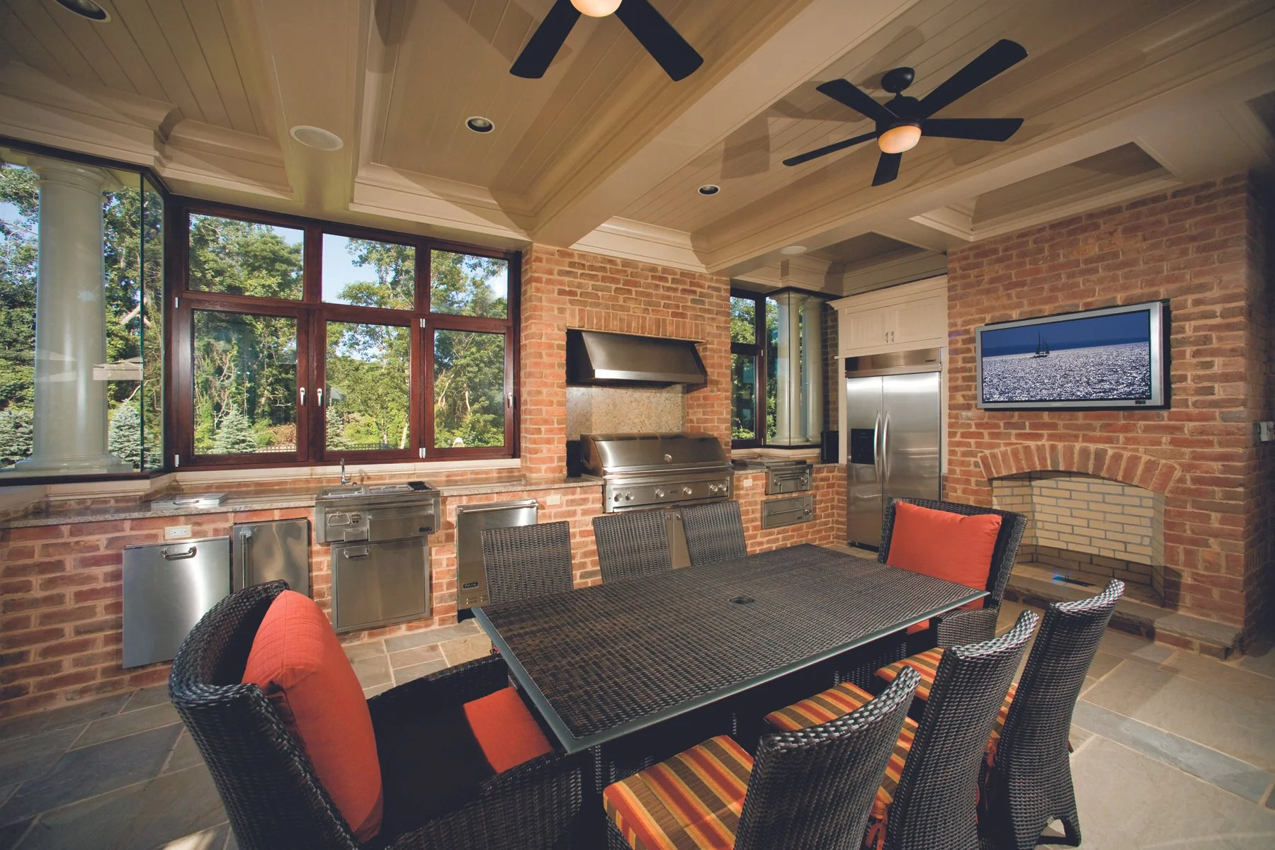 Indoor outdoor kitchen with brick walls, large window, stainless steel grill, refrigerator, TV, and a dining table with six chairs and orange cushions.