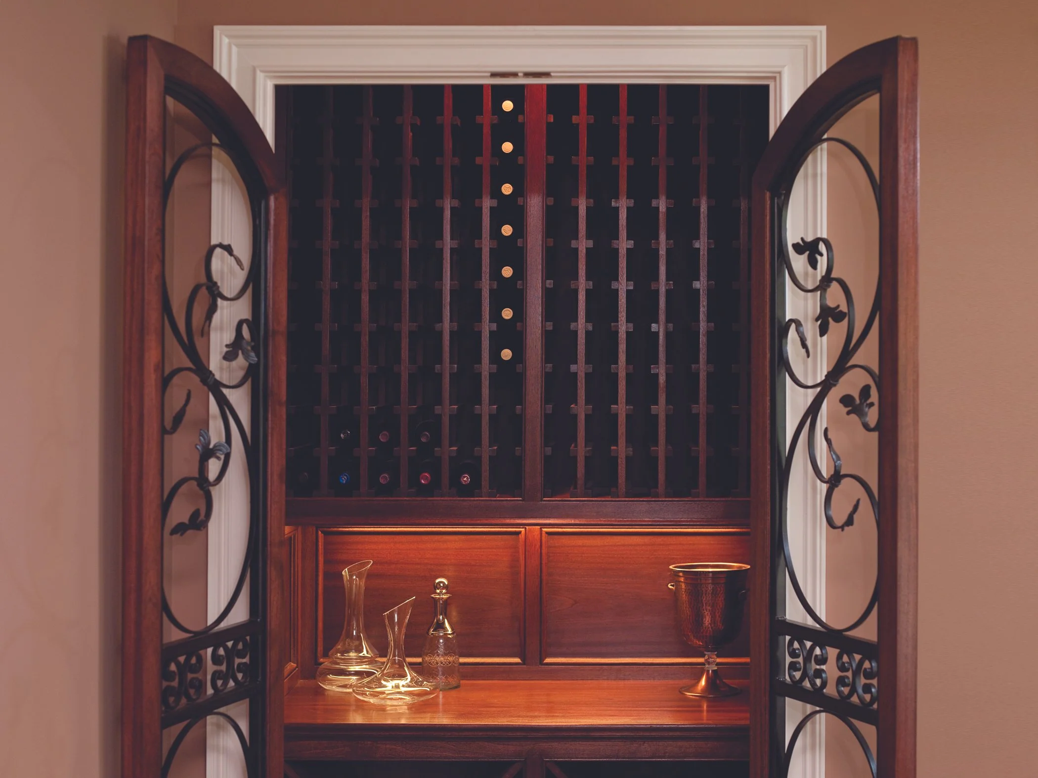 A decorative wine cellar with wooden racks, glass decanters, and a wine glass, framed by wrought iron doors with floral designs.