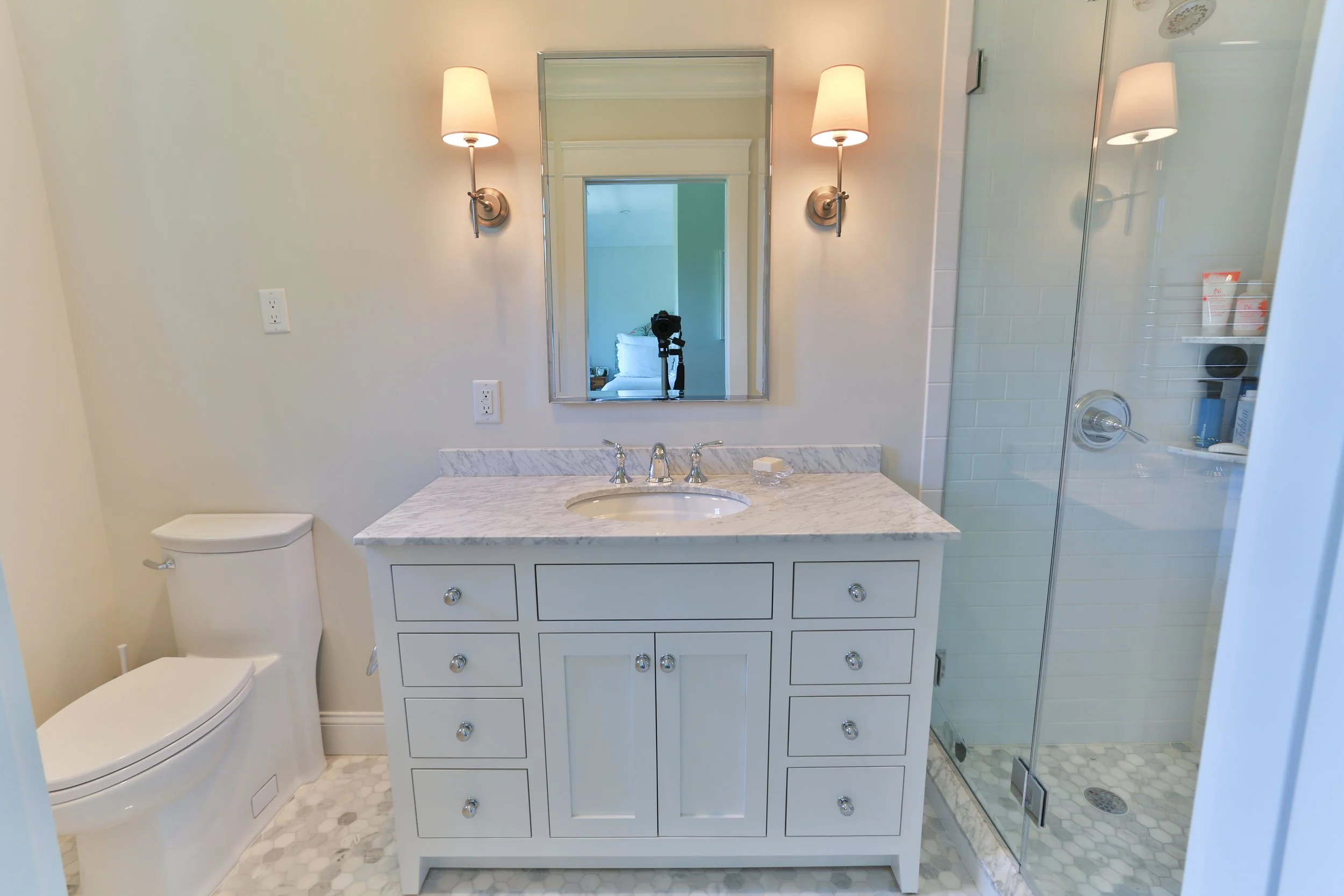 Bathroom with a white vanity with a marble top, a mirror, beige wall sconces, a toilet, and a glass shower enclosure with white tile.