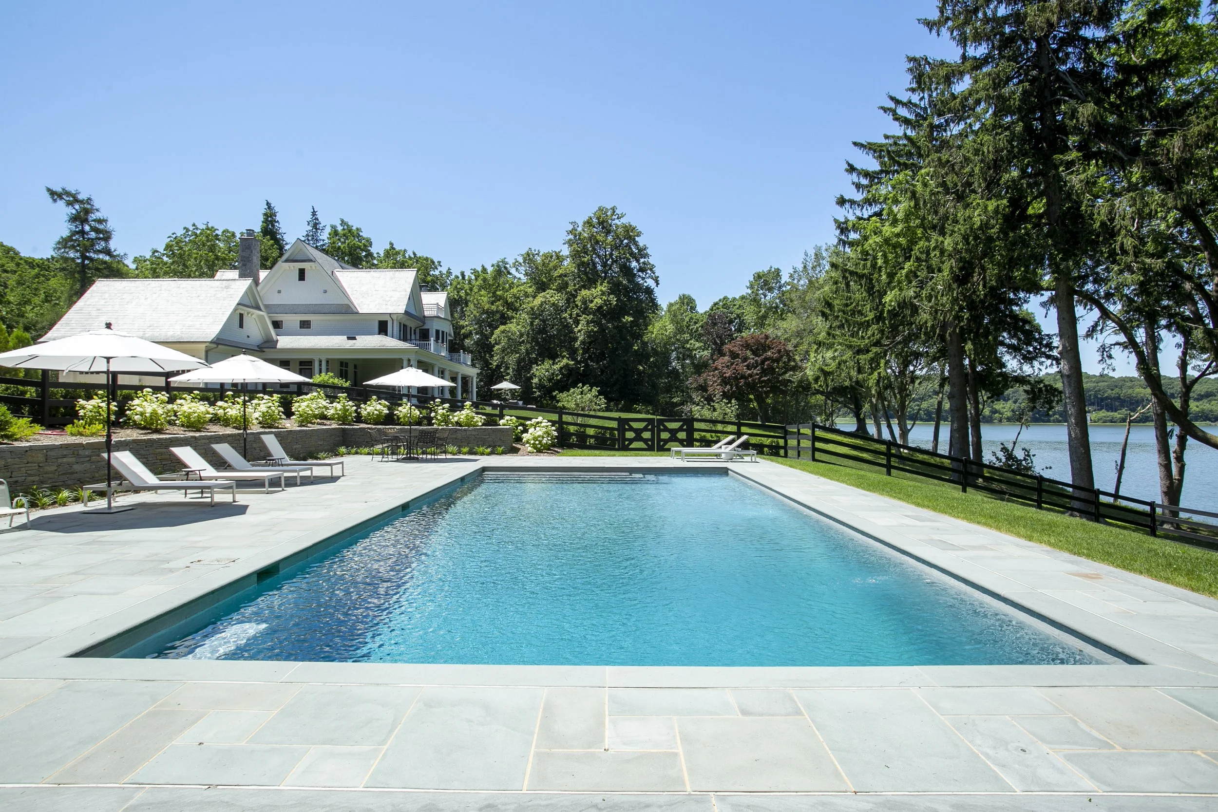 A swimming pool outdoors with lounge chairs and umbrellas, a large house in the background, trees, and a body of water on the right.