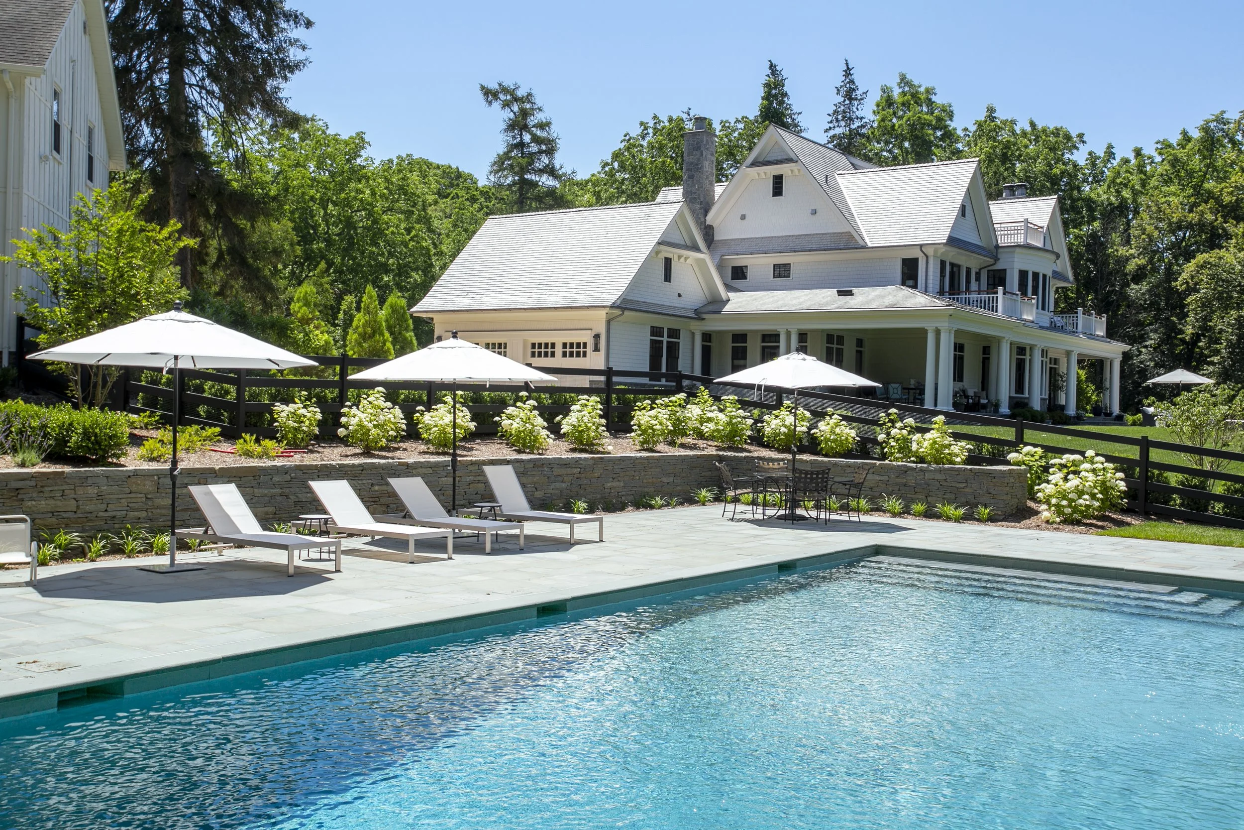 Residential house with a swimming pool, lounge chairs with umbrellas, and landscaped garden with flowering bushes.