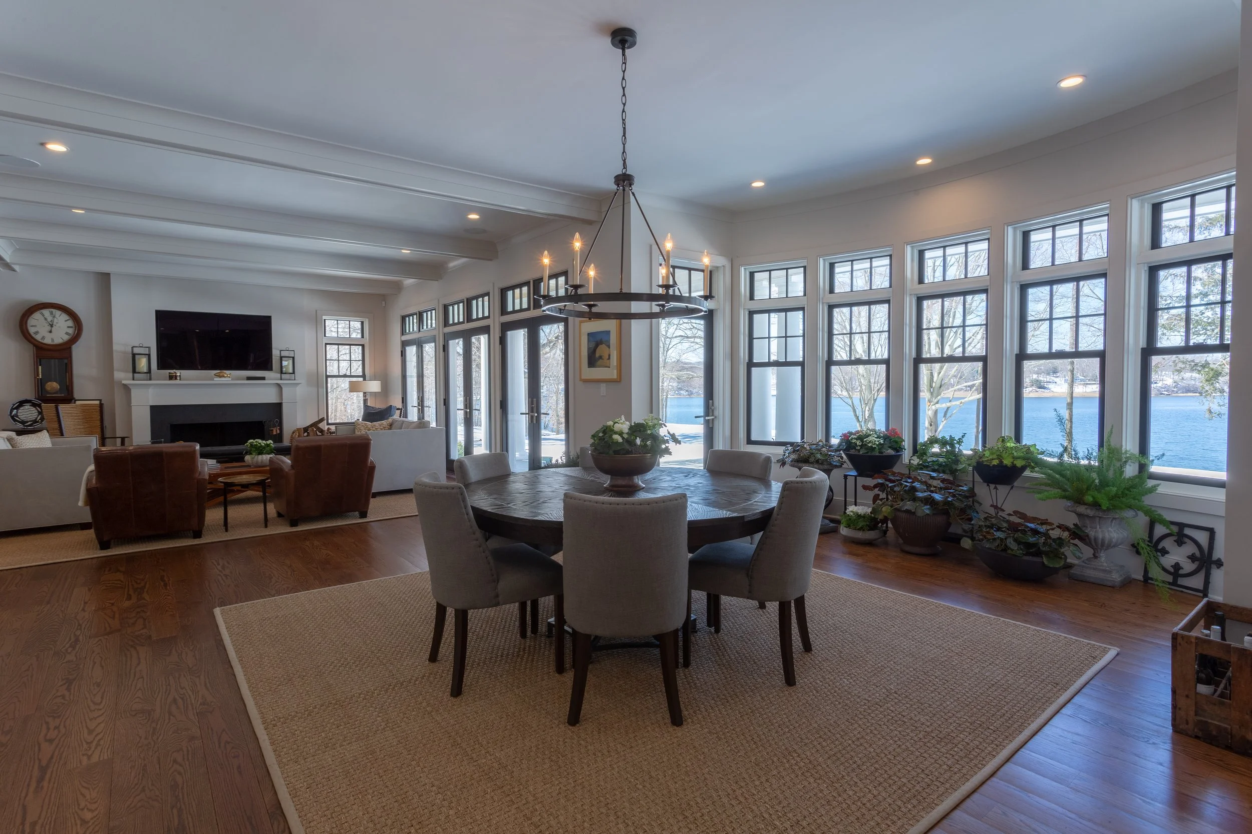Spacious living room with large bay windows overlooking water, open floor plan, round dining table with eight chairs, and comfortable seating area with sofas and armchairs near a fireplace.