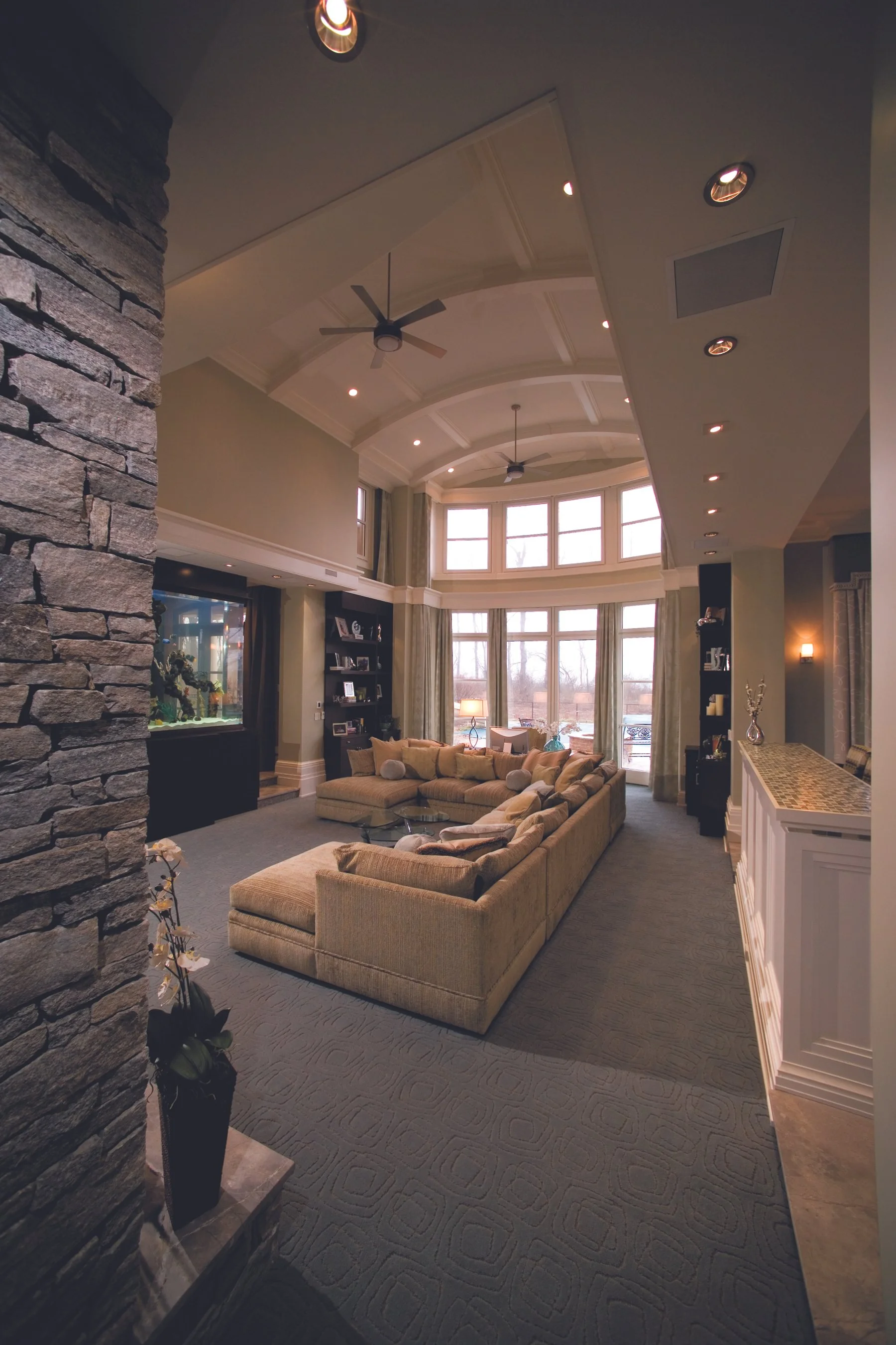 Spacious living room with a high vaulted ceiling, large bay windows, a beige sectional sofa, built-in bookshelves, a stone accent wall, and ceiling fans.