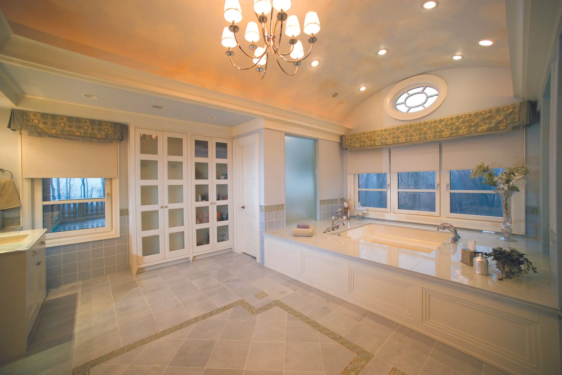A spacious bathroom with a large bathtub, multiple windows, a chandelier, and built-in cabinetry.