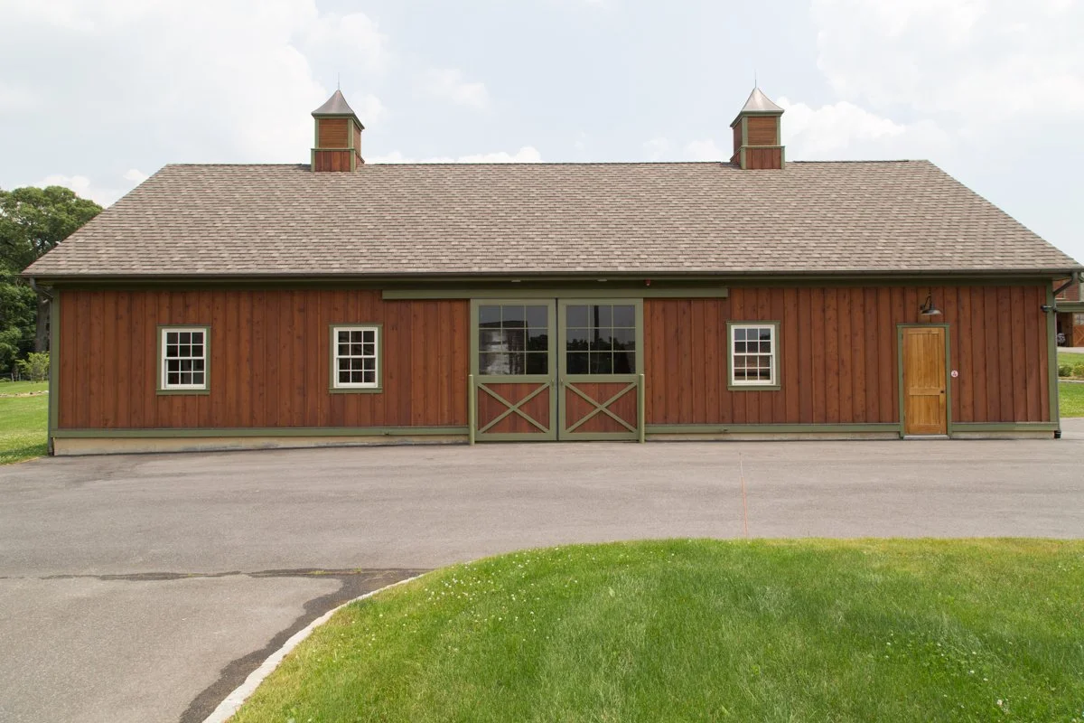 A wooden barn with a shingled roof, two small chimneys, and three windows, with a sliding barn door in the center and a side door on the right, situated on a paved area with grass in the foreground.