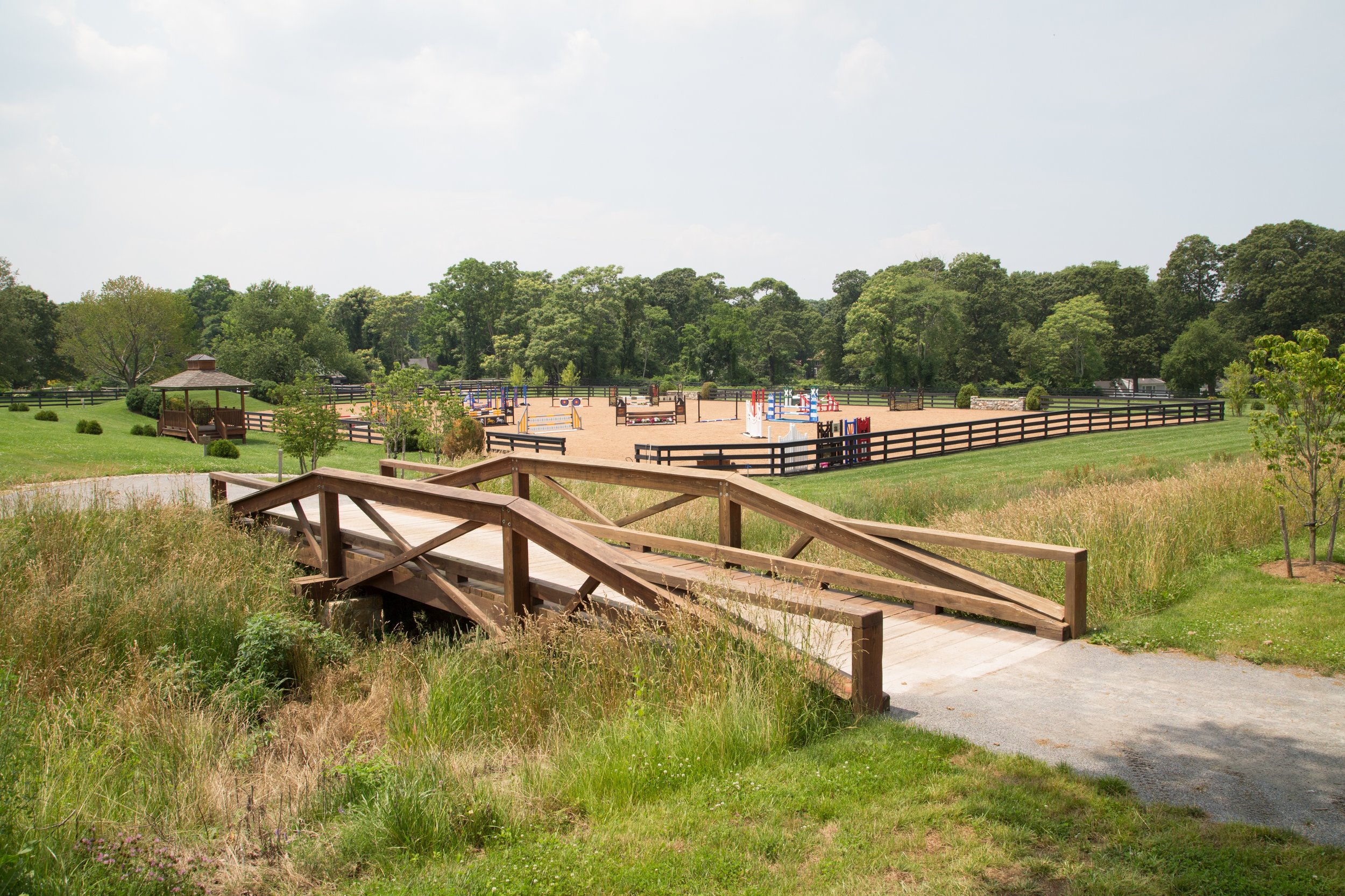 A horse riding arena with colorful jumps, surrounded by green trees and grass, and a wooden bridge in the foreground.