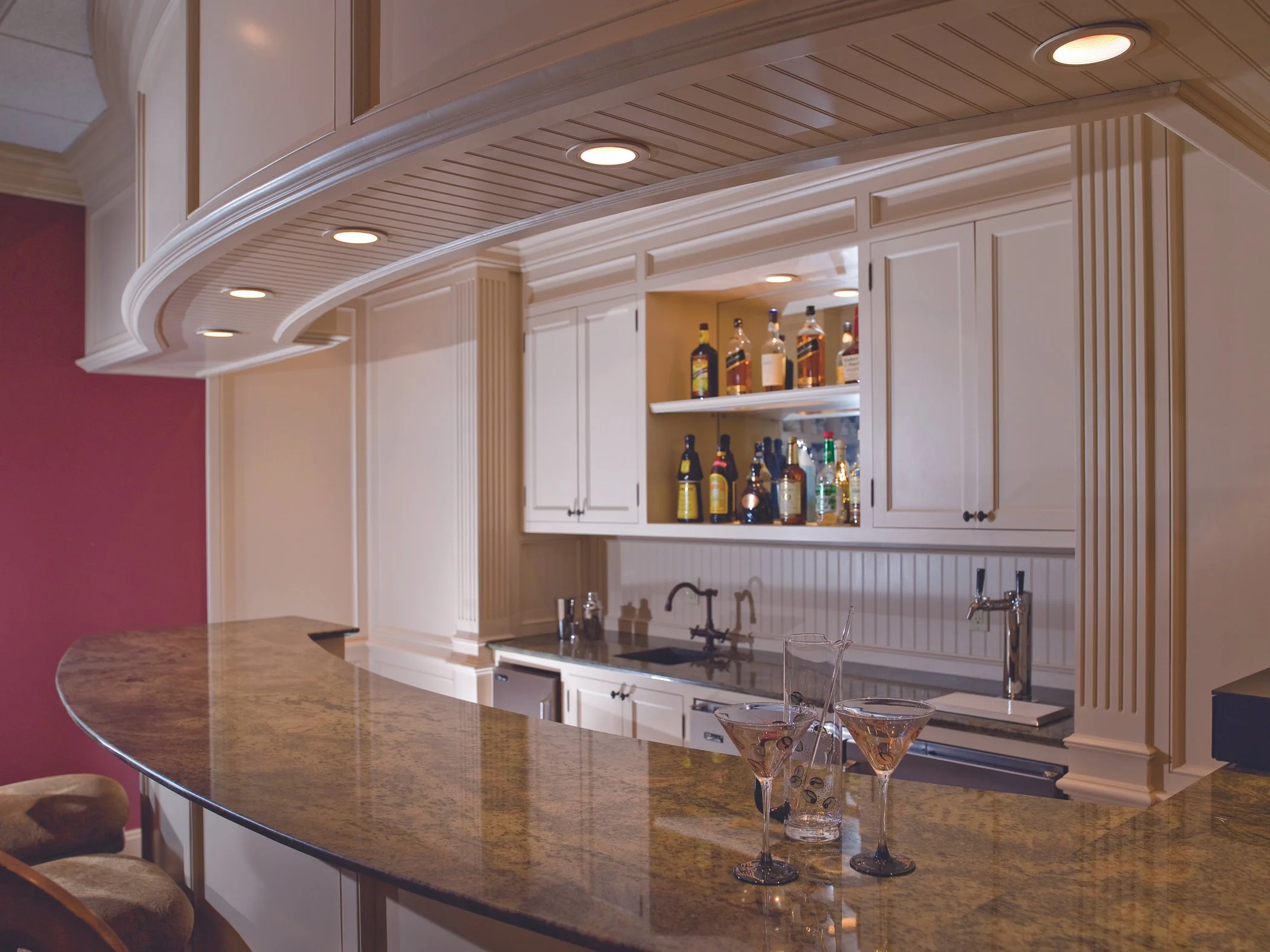 Interior view of a home bar with a curved granite countertop, white cabinets, and a selection of liquor bottles on open shelves, with two martini glasses on the countertop.