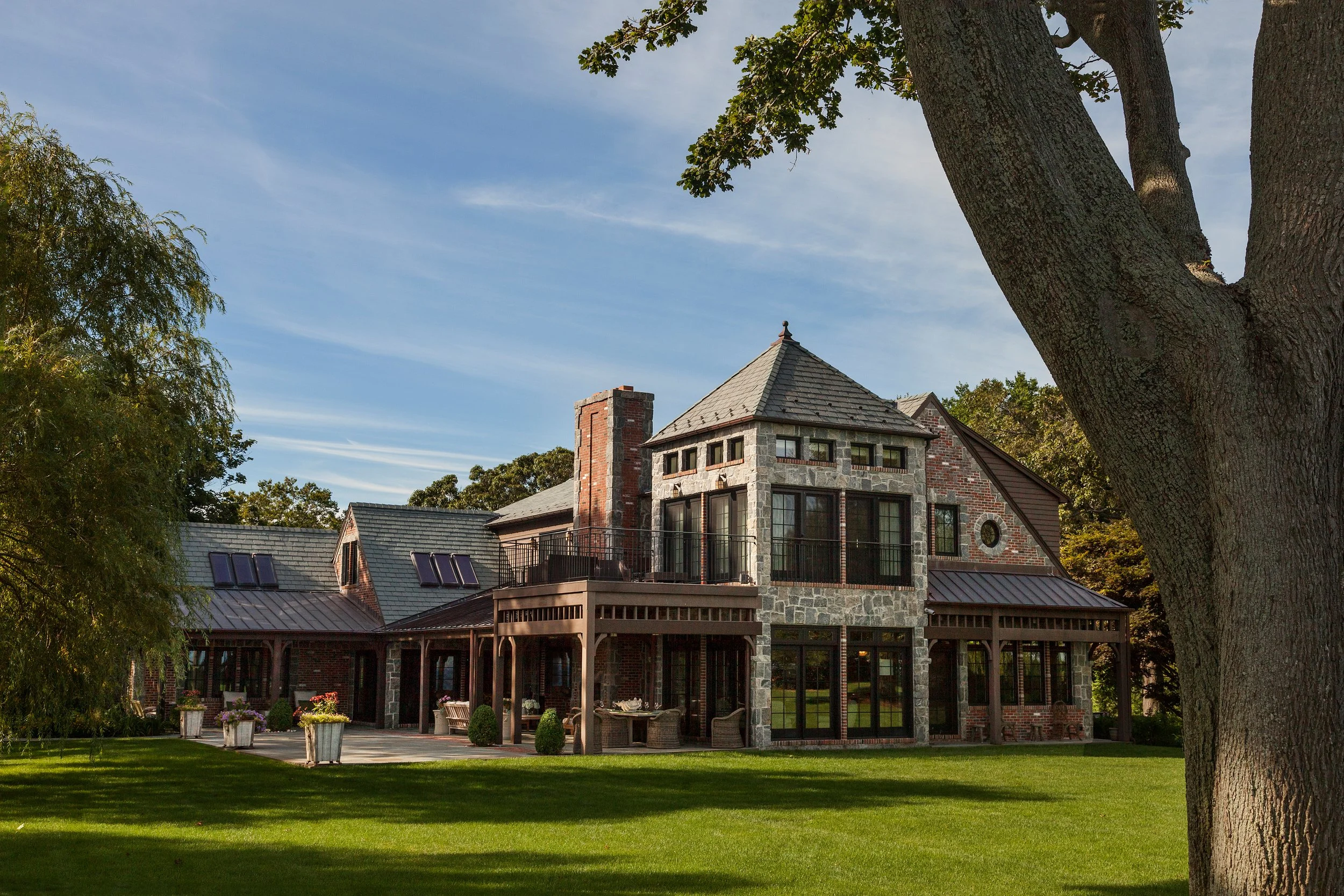 A large house with a stone and brick exterior, multiple rooflines including a tower-like section, large windows, a chimney, and an outdoor patio with chairs and potted plants, set against a bright blue sky and surrounded by green grass and trees.