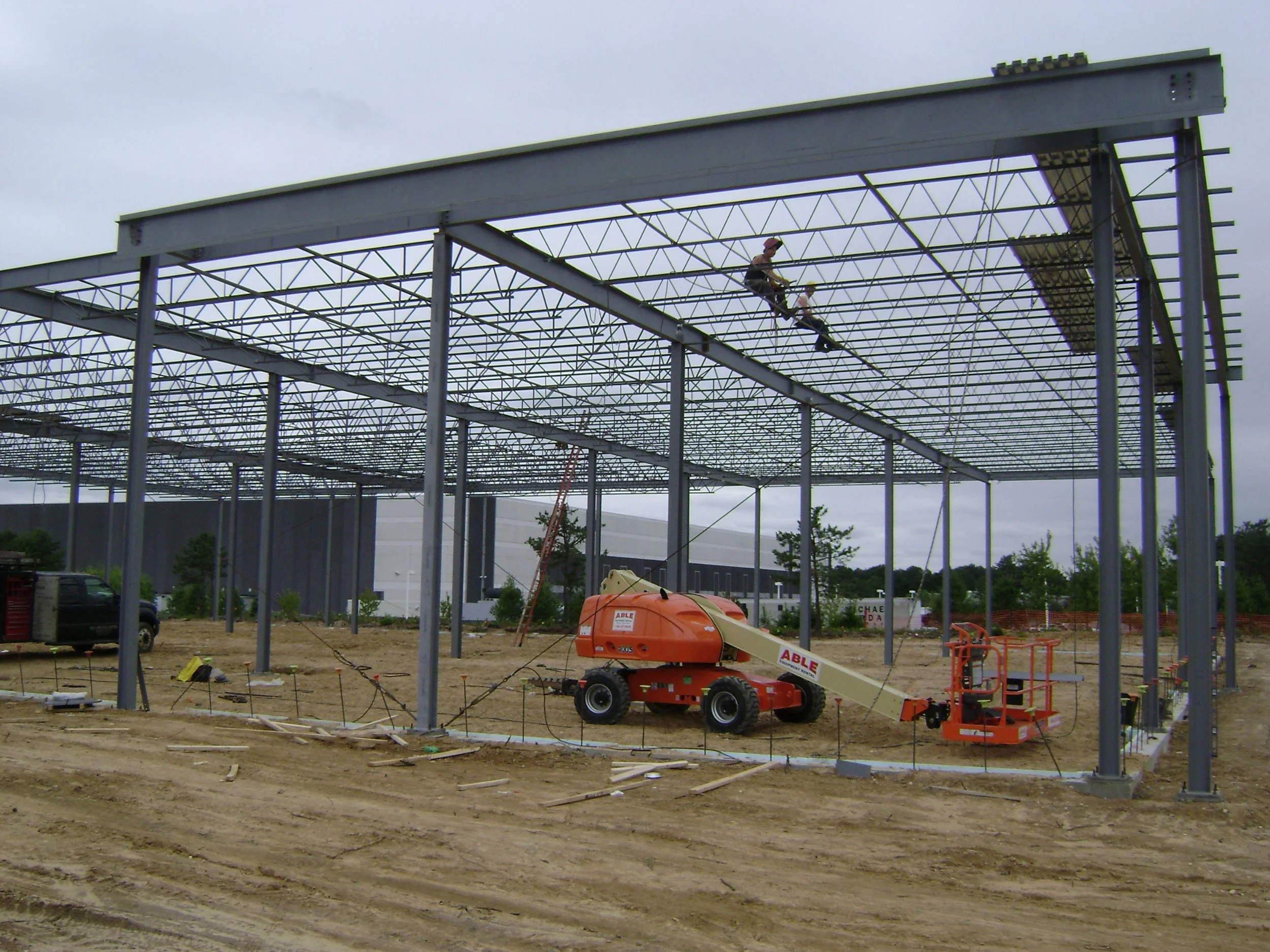 Construction workers build a metal framework for a structure, with one worker on scaffolding and others operating cranes on a dirt site.