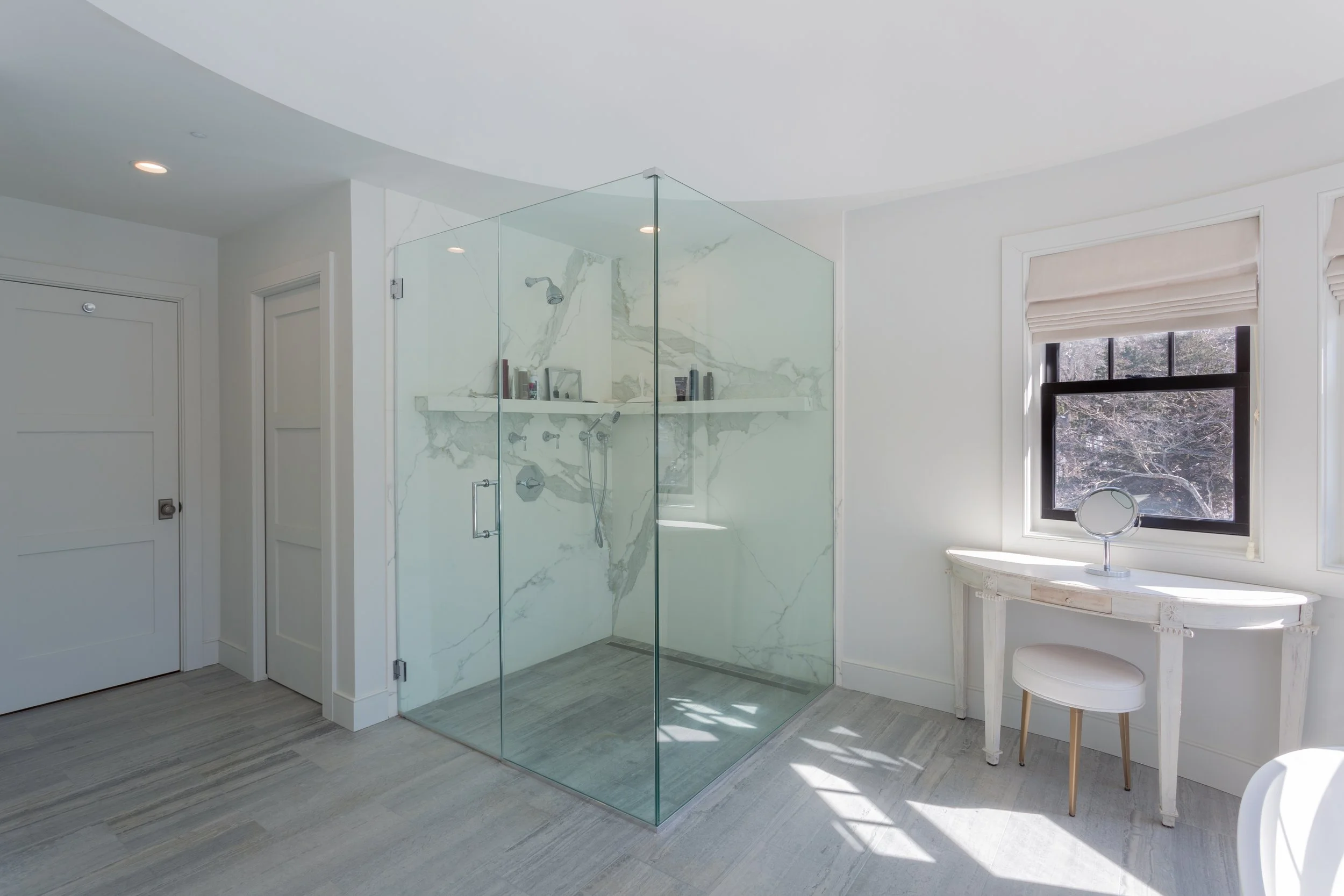 A modern bathroom with a walk-in glass shower, white marble walls, a window with a Roman shade, and a small white dressing table with a mirror