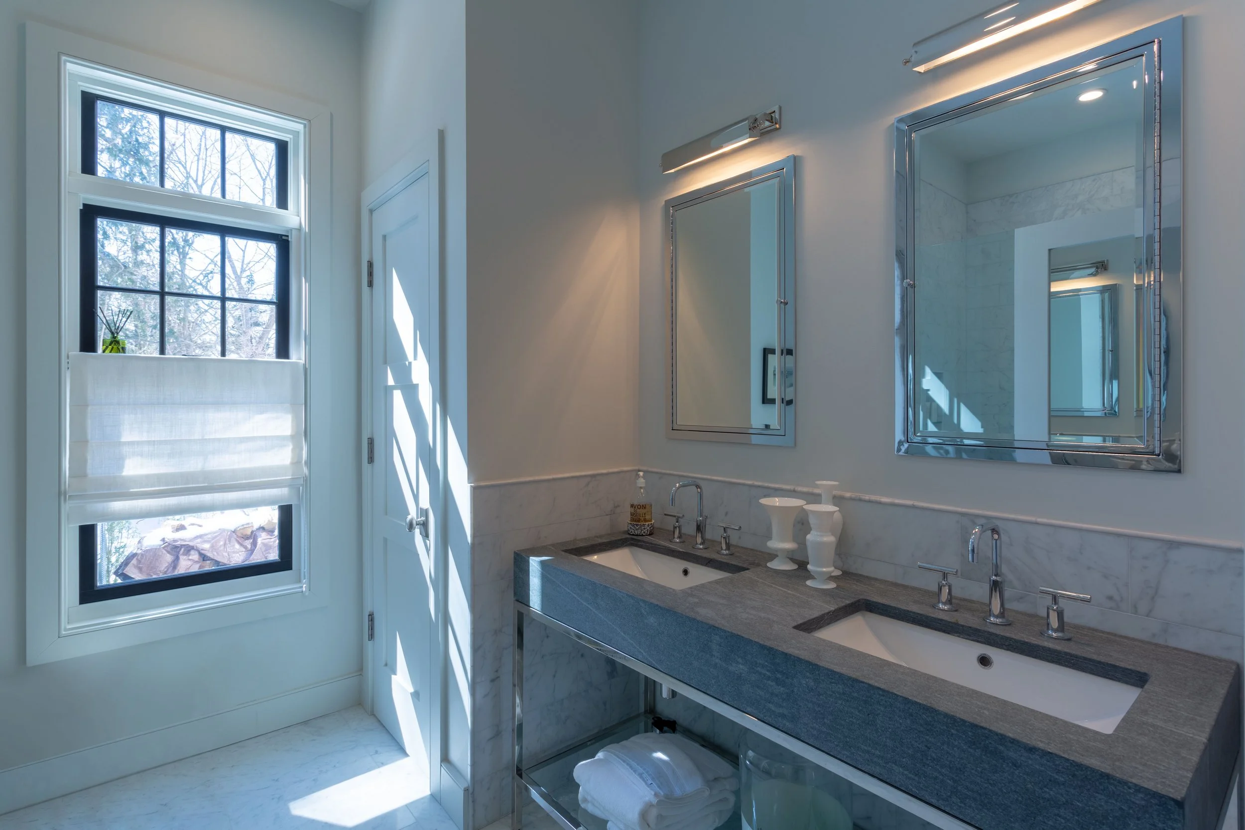 Bathroom with double sinks, marble countertops, and mirrors, illuminated by modern wall lights. A window with a partly rolled-up white blind lets in natural light. Decorative vases on the counter.