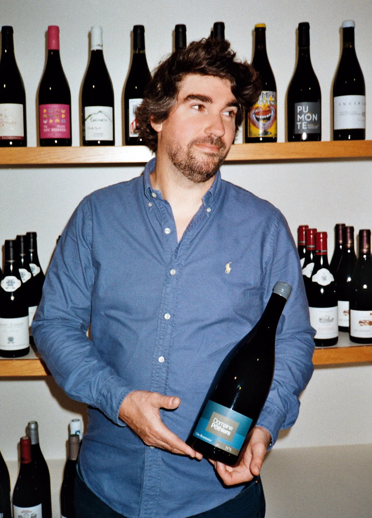 Kévin Defarges, caviste tenant une bouteille de vin du Romain Paris domaine des Pothiers dans une cave à vin, entouré de nombreuses autres bouteilles de vin sur des étagères.