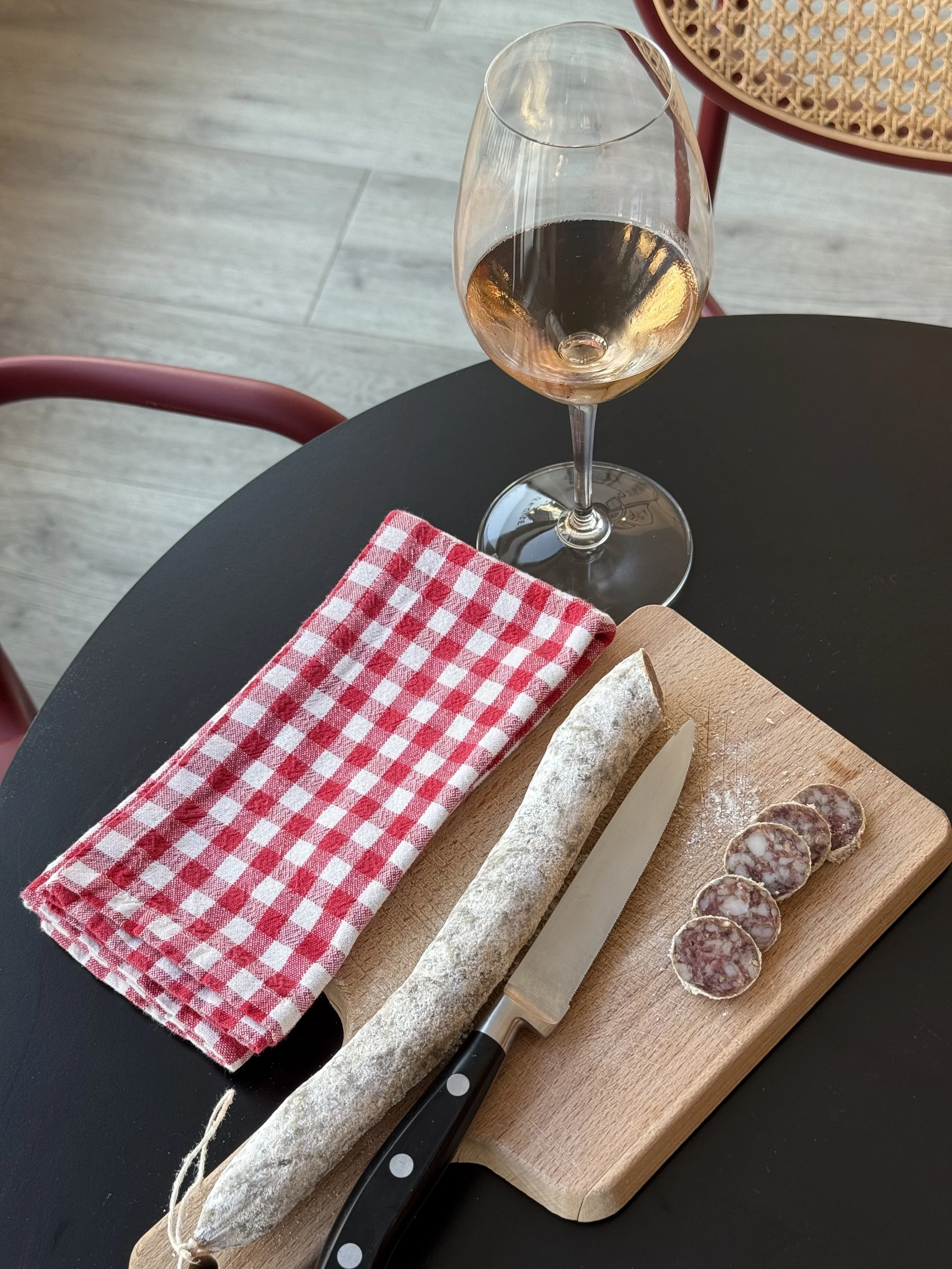 Verre de vin rosé sur une table noire avec une planche à découper, un couteau, un saucisson, et une serviette à carreaux rouges et blancs esprit guinguette.