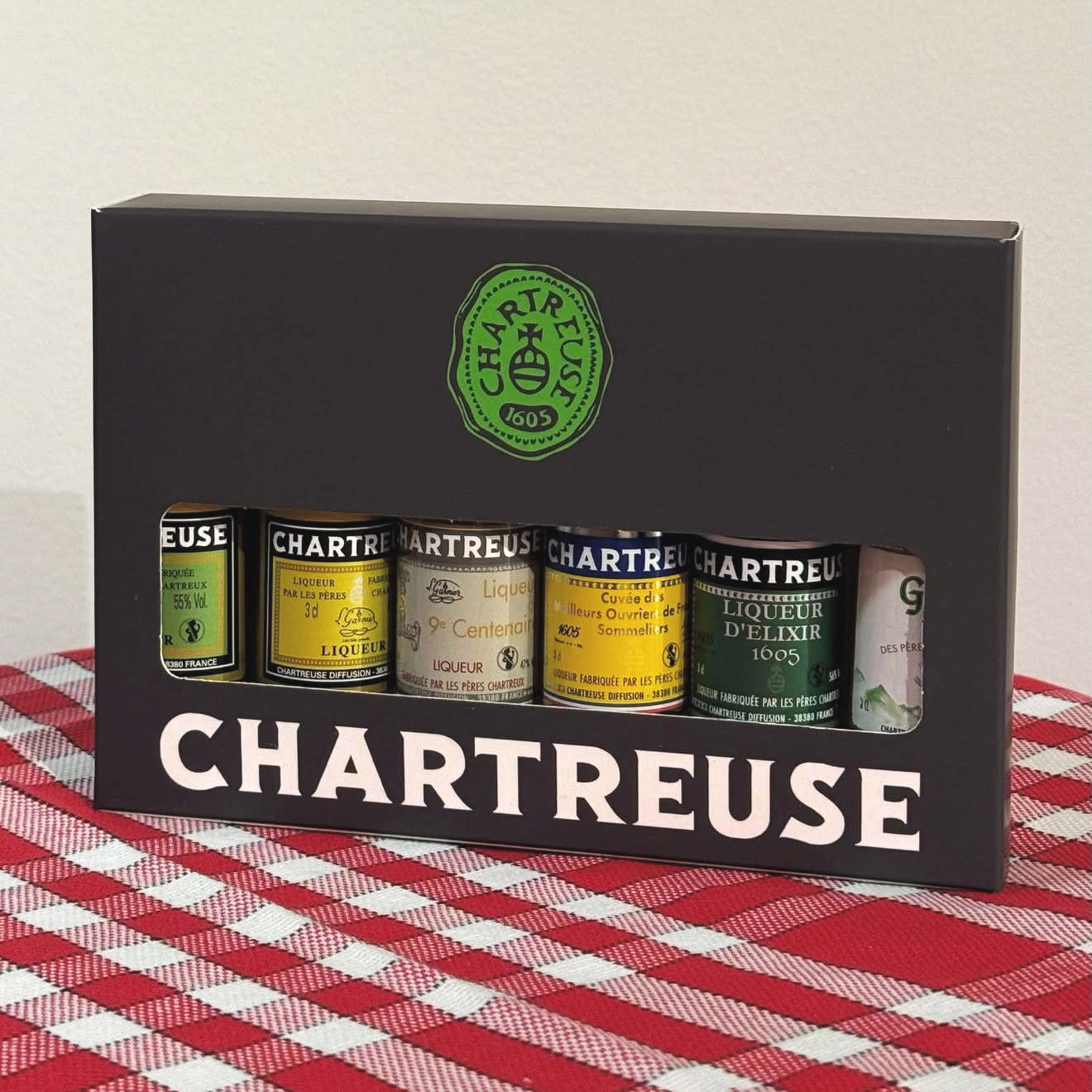 Coffret de cinq bouteilles de liqueur Chartreuse de différentes variétés, présenté dans une boîte noire avec une fenêtre en plastique. Le coffret est posé sur une table à carreaux rouge et blanc.
